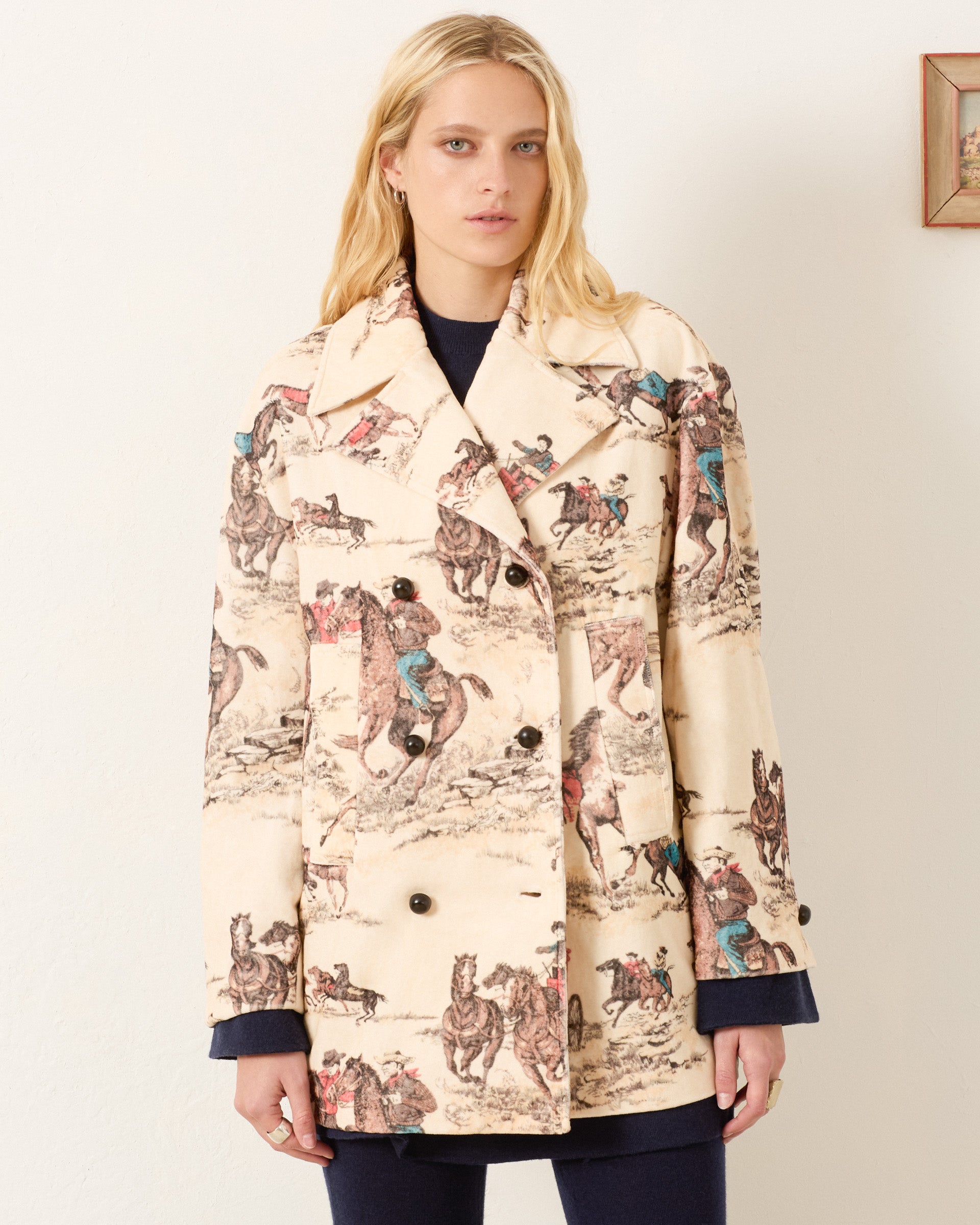 A woman stands against a plain wall wearing the Rivoli Canyon Cowboy Peacoat, a cream-colored cotton-velvet, double-breasted coat printed with striking horse and rider illustrations.
