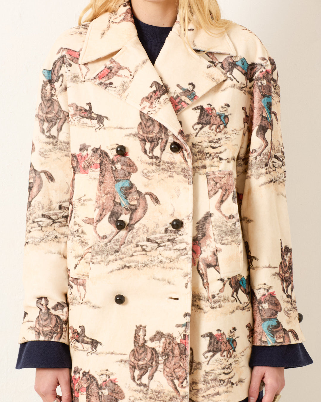 Someone wears the Rivoli Canyon Cowboy Peacoat, a light-toned cotton-velvet Western coat adorned with a detailed horse and rider action print.