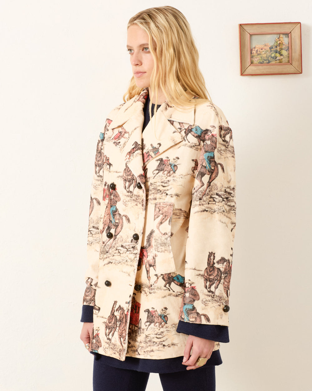 A woman wearing the Rivoli Canyon Cowboy Peacoat, a cream-colored cotton-velvet coat with a horse and rider print, stands indoors beside a small framed painting on the wall.