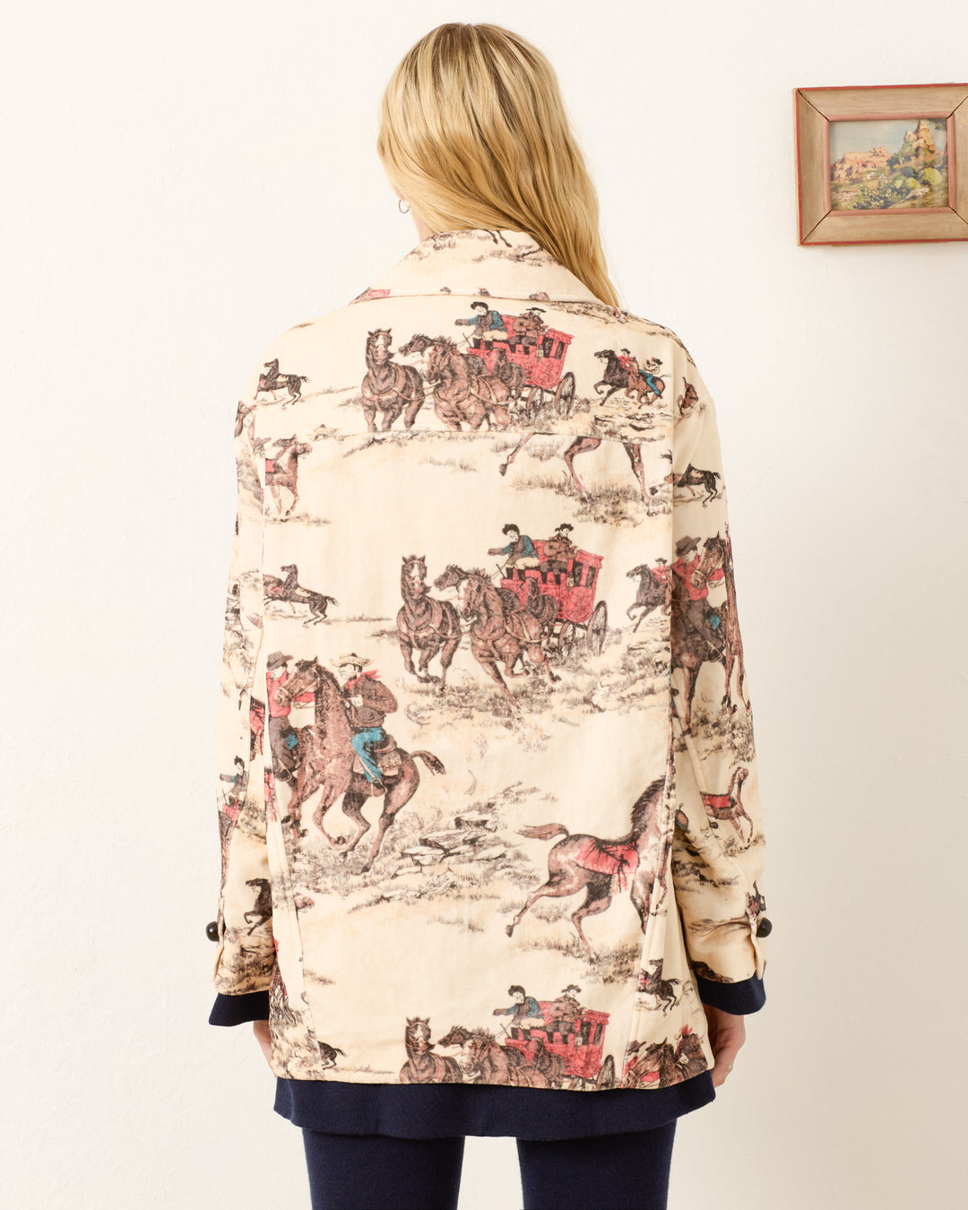 A person with long blonde hair, seen from behind, wears the Rivoli Canyon Cowboy Peacoat—a cream cotton-velvet Western coat with a vintage horse and stagecoach print. To the right, a small framed painting hangs on the wall.