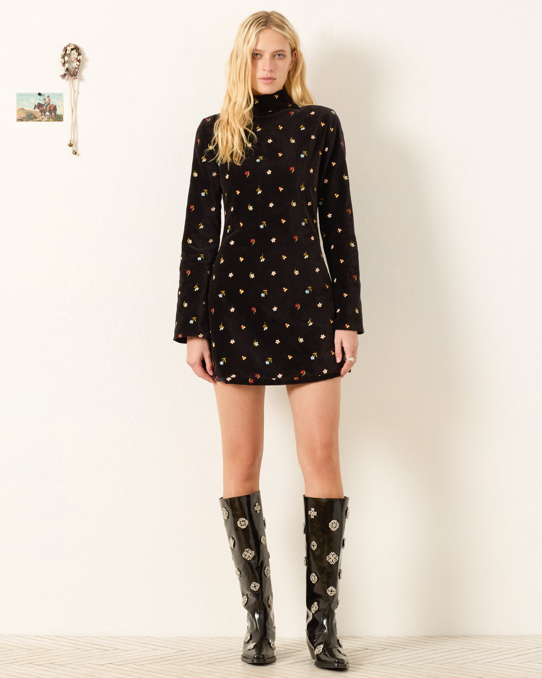A woman faces forward in the Romy Onyx Bouquet Dress—a black cotton-velvet mini dress with a small colorful print and long flared sleeves—paired with knee-high black boots, posing against a simple white wall.