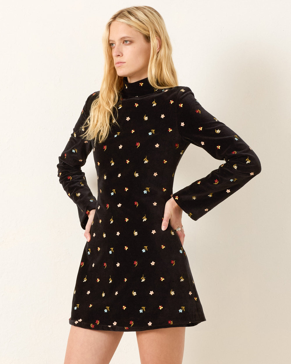 A woman wears the Romy Onyx Bouquet Dress, a black cotton-velvet mini dress with long flared sleeves, a high neckline, and small colorful floral embroidery as she stands against a plain light background.