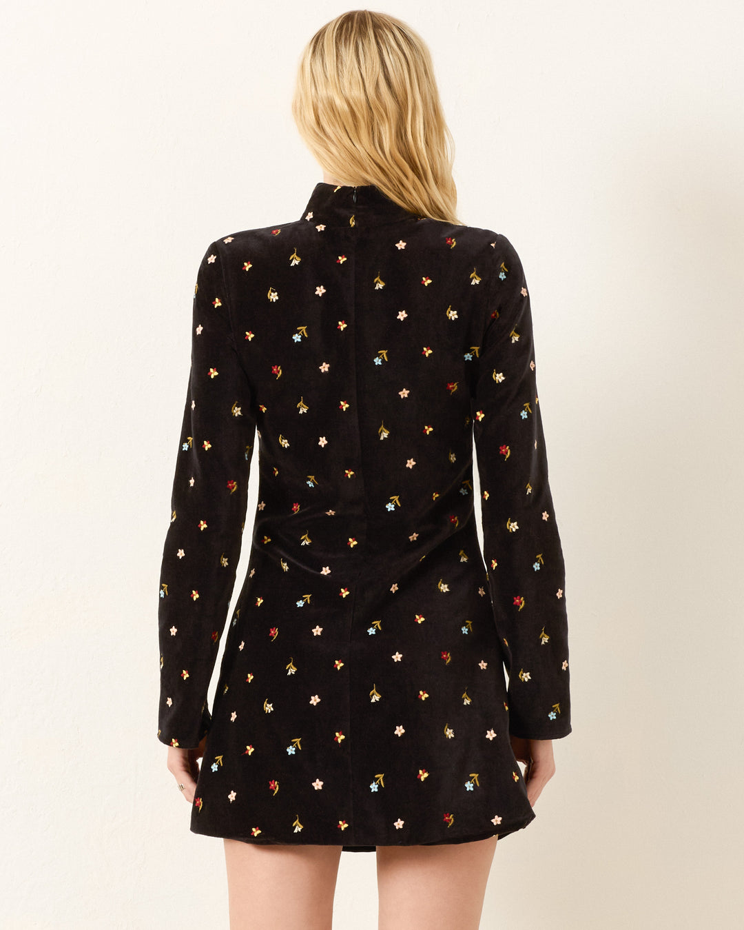 A woman stands facing away, wearing the Romy Onyx Bouquet Dress—a black cotton-velvet mini dress with a high collar, long flared sleeves, and small colorful floral patterns.