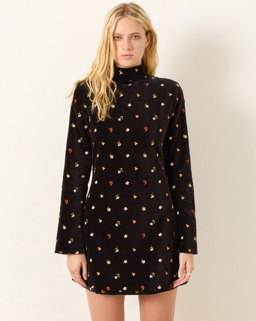 A woman wears the Romy Onyx Bouquet Dress—a long-sleeve black turtleneck dress with a multicolored small floral print—standing against a plain light background.
