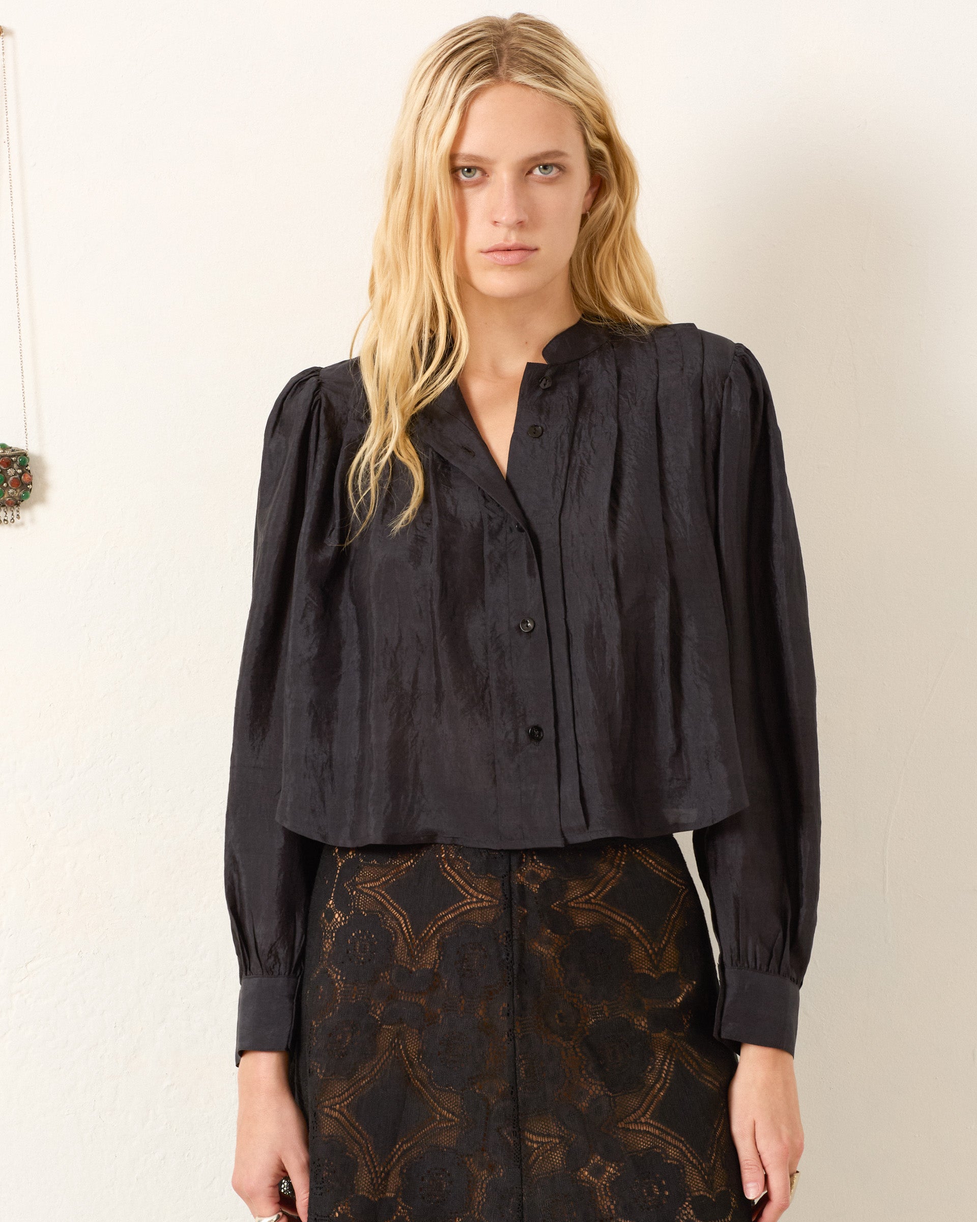 A woman with long blonde hair wears the Ryle Onyx Silk Shirt and a patterned high-waisted skirt, standing against a plain light-colored wall.