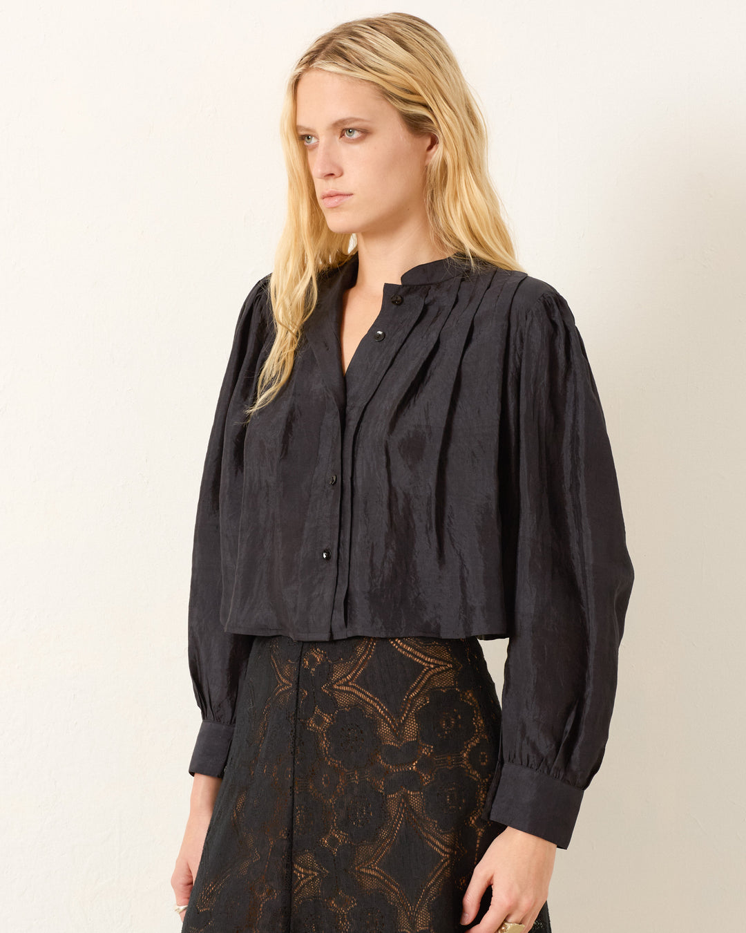 A woman with long blonde hair wears the Ryle Onyx Silk Shirt, a black Victorian-style blouse with buttons and a high collar, paired with a black lace skirt, against a plain light background.