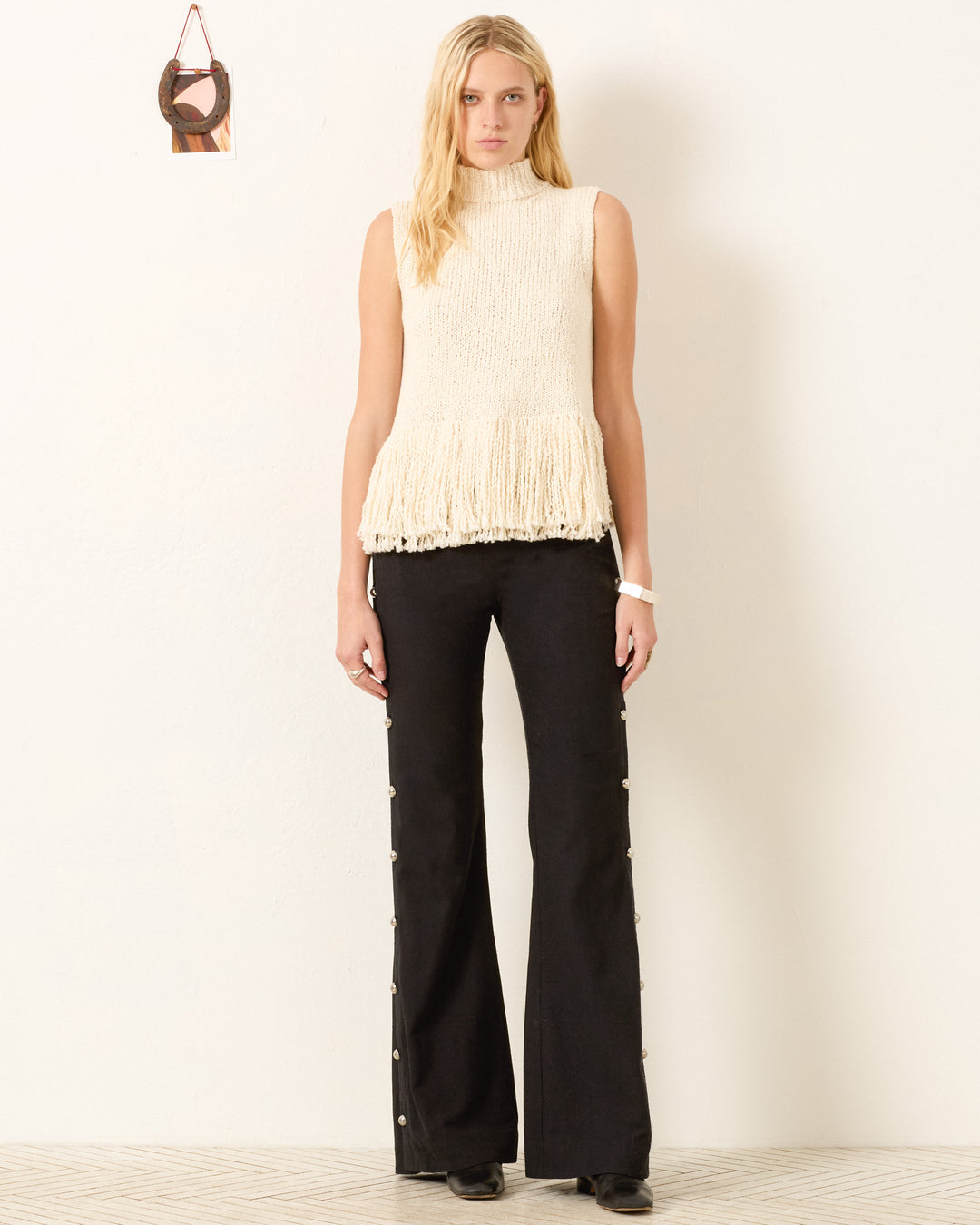 A woman stands against a white wall wearing the artisan-made Sonia Snow Fringe Top—a sleeveless, cream textured top with a fringe hem—paired with black wide-leg pants, black shoes, and a small bag hanging on the wall.