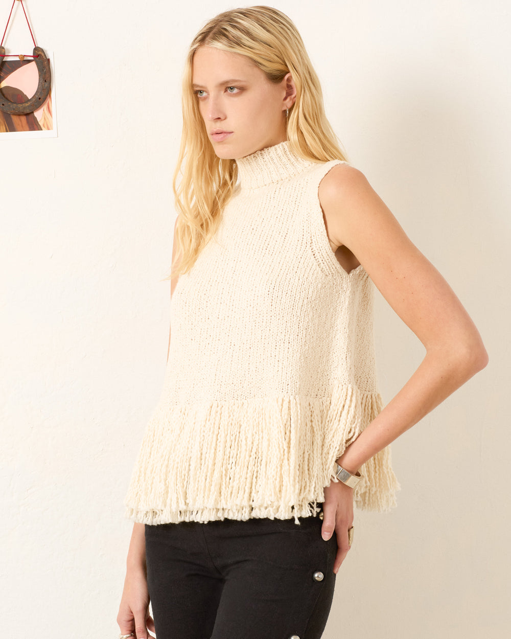 A woman with long blonde hair wears the Sonia Snow Fringe Top, an artisan-made, hand-knitted cream mock-neck with a fringed hem, paired with black pants against a plain white wall.