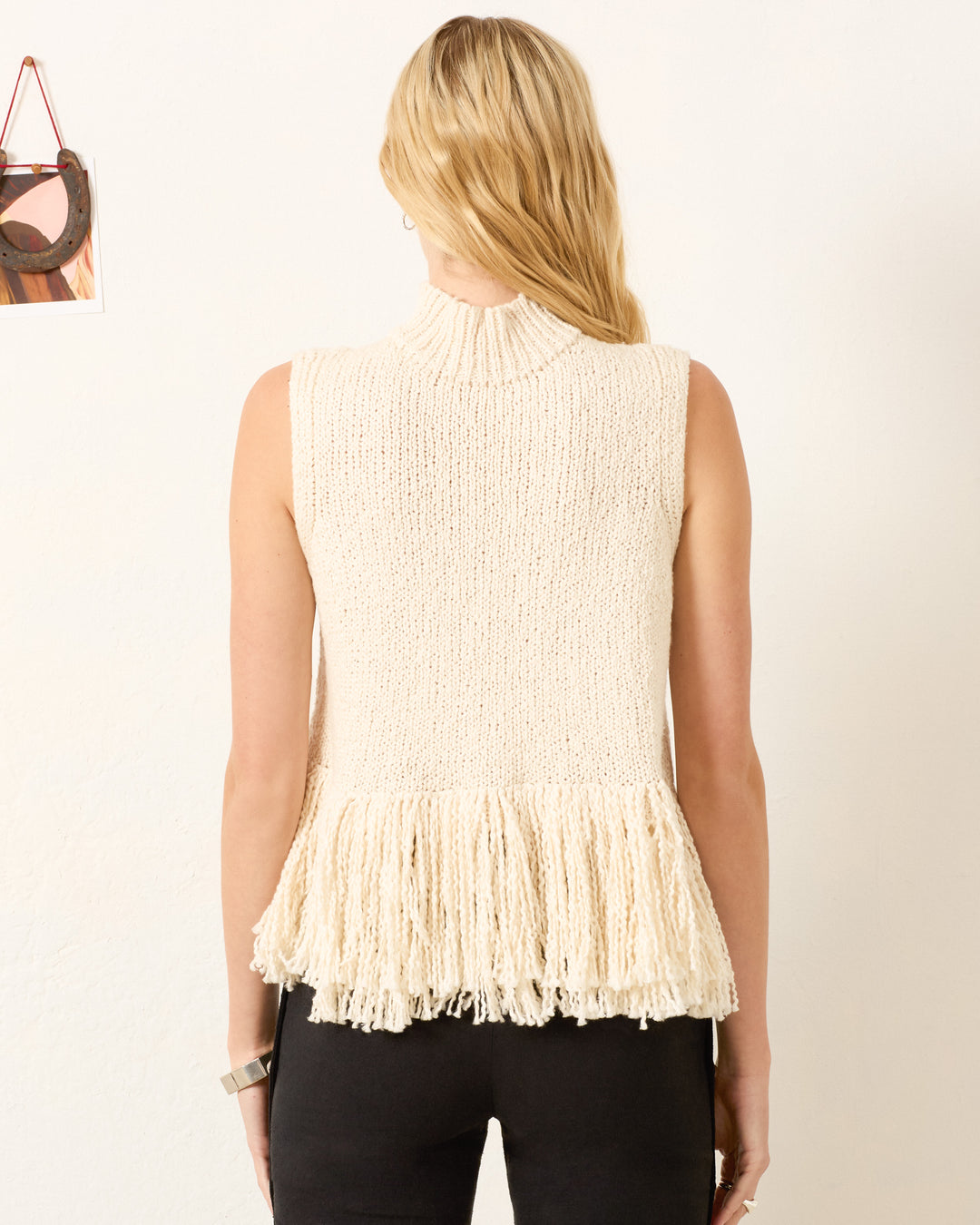 A woman with long blonde hair models the artisan-made Sonia Snow Fringe Top, a sleeveless cream knit design with fringe at the hem, standing with her back to the camera against a plain wall.