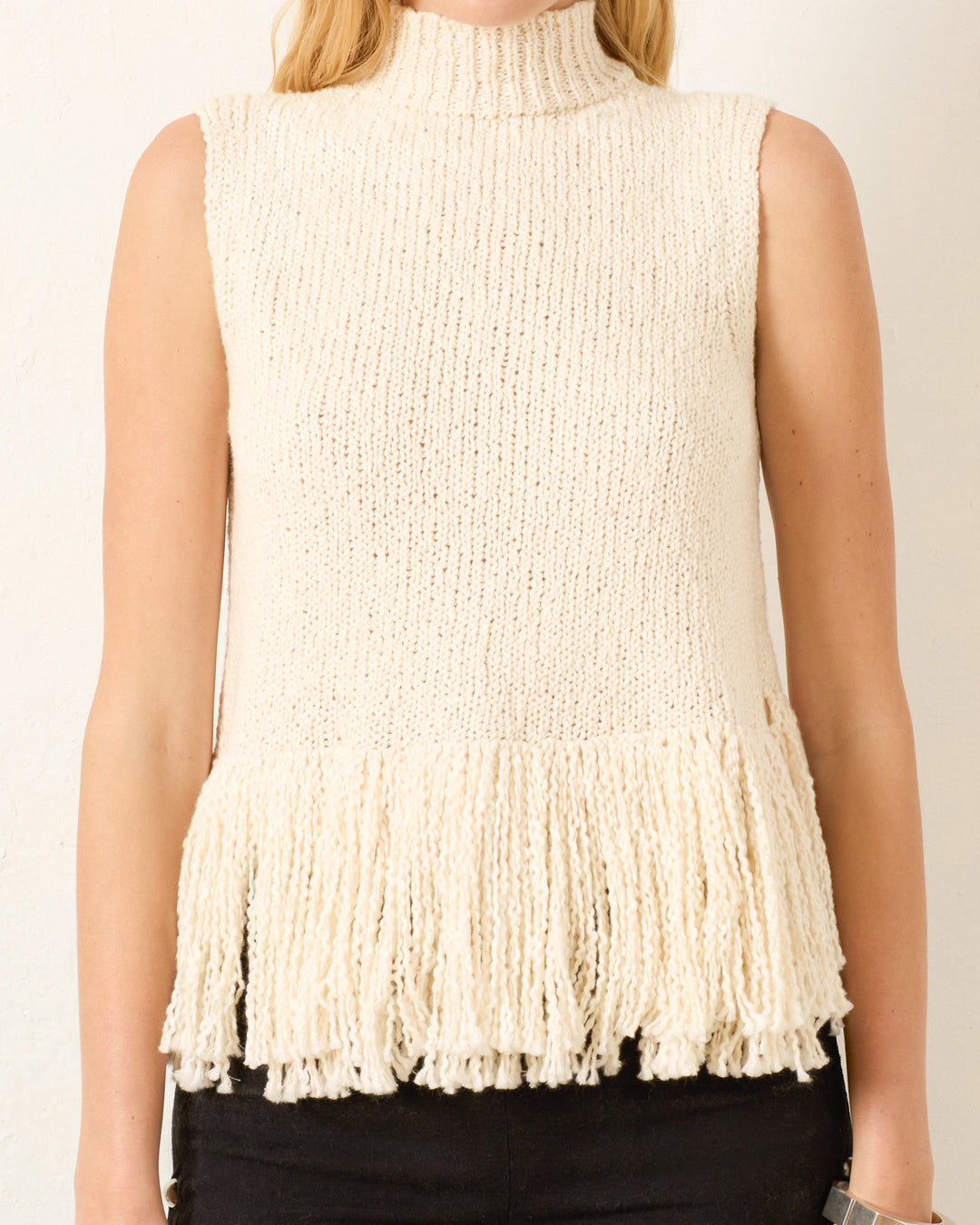 A blonde-haired model wears the Sonia Snow Fringe Top—a sleeveless, cream hand-knitted mock-neck top with a fringed hem, crafted from organic cotton.