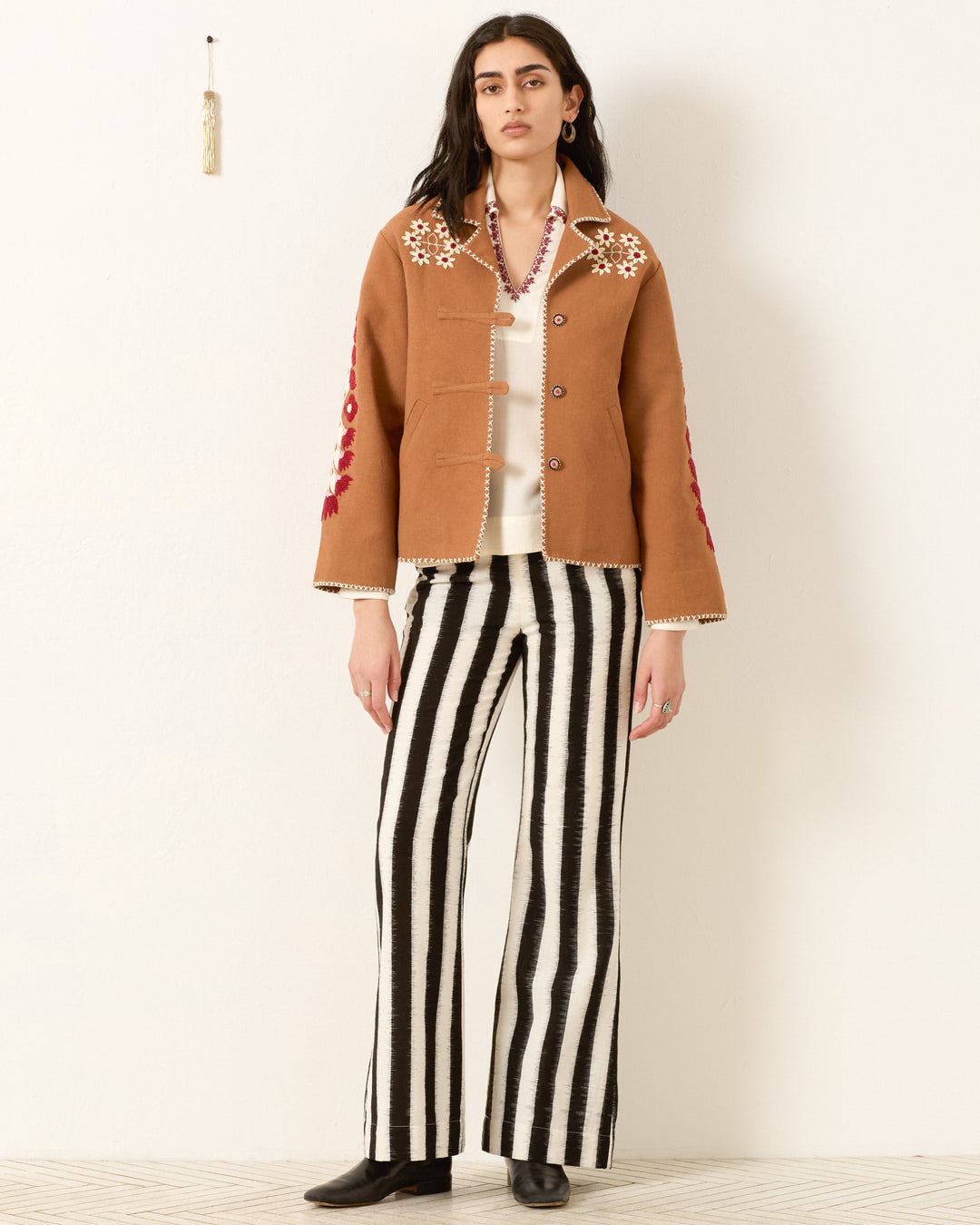 Wearing the Western Cactus Bloom Jacket with floral embroidery, a white shirt, black and white striped pants, black shoes, and a necklace, a person stands against a white wall.