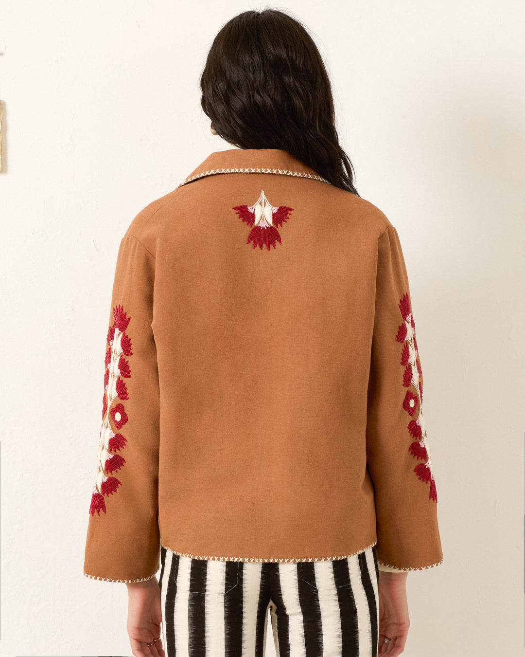 A person with long dark hair, seen from behind, wears the Western Cactus Bloom Jacket in tan with red and white embroidery on the sleeves and back, paired with black and white striped pants.