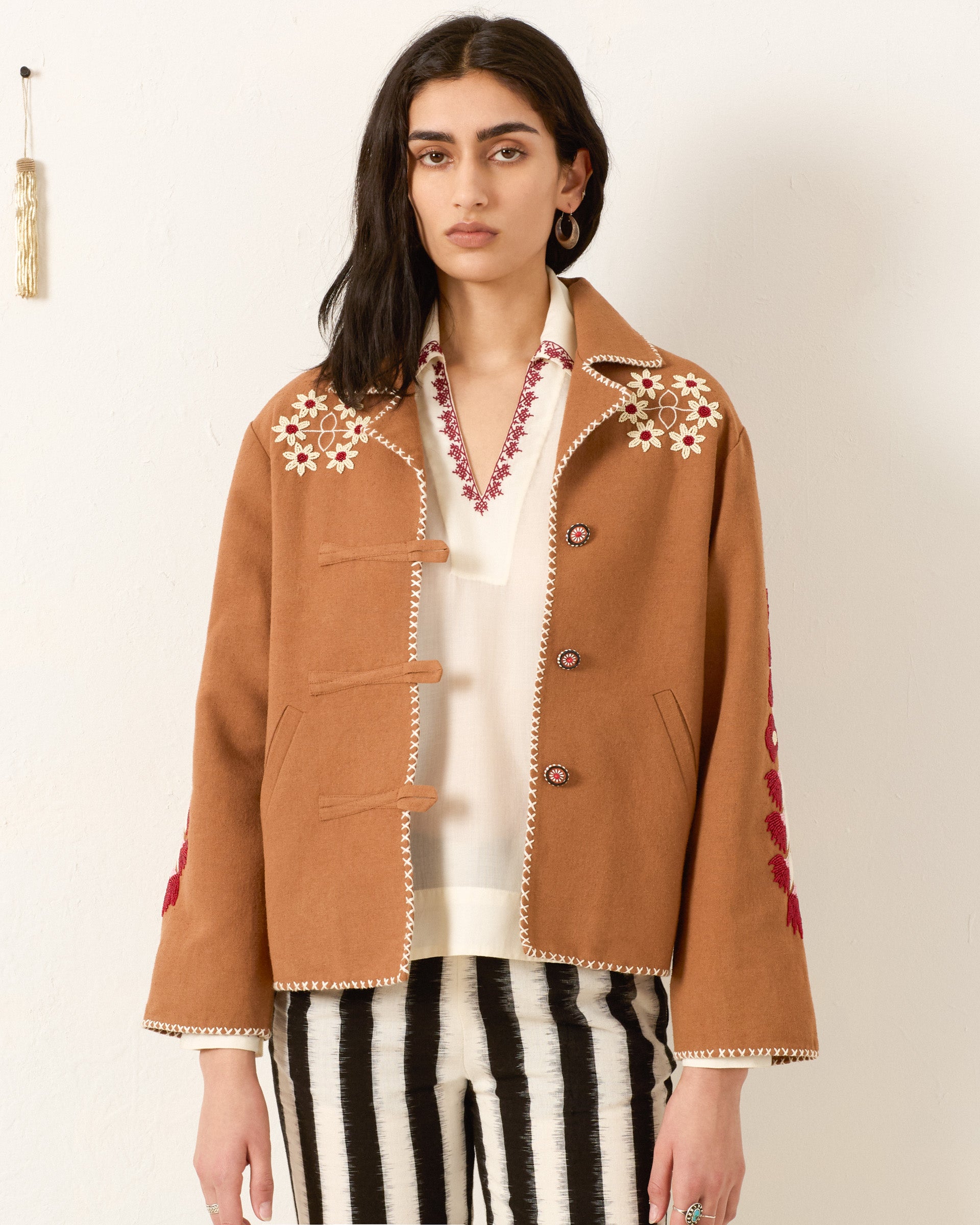 A woman stands against a white wall wearing the Western Cactus Bloom Jacket with hand-beaded floral motif, paired with a white shirt featuring red details and black-and-white striped pants.