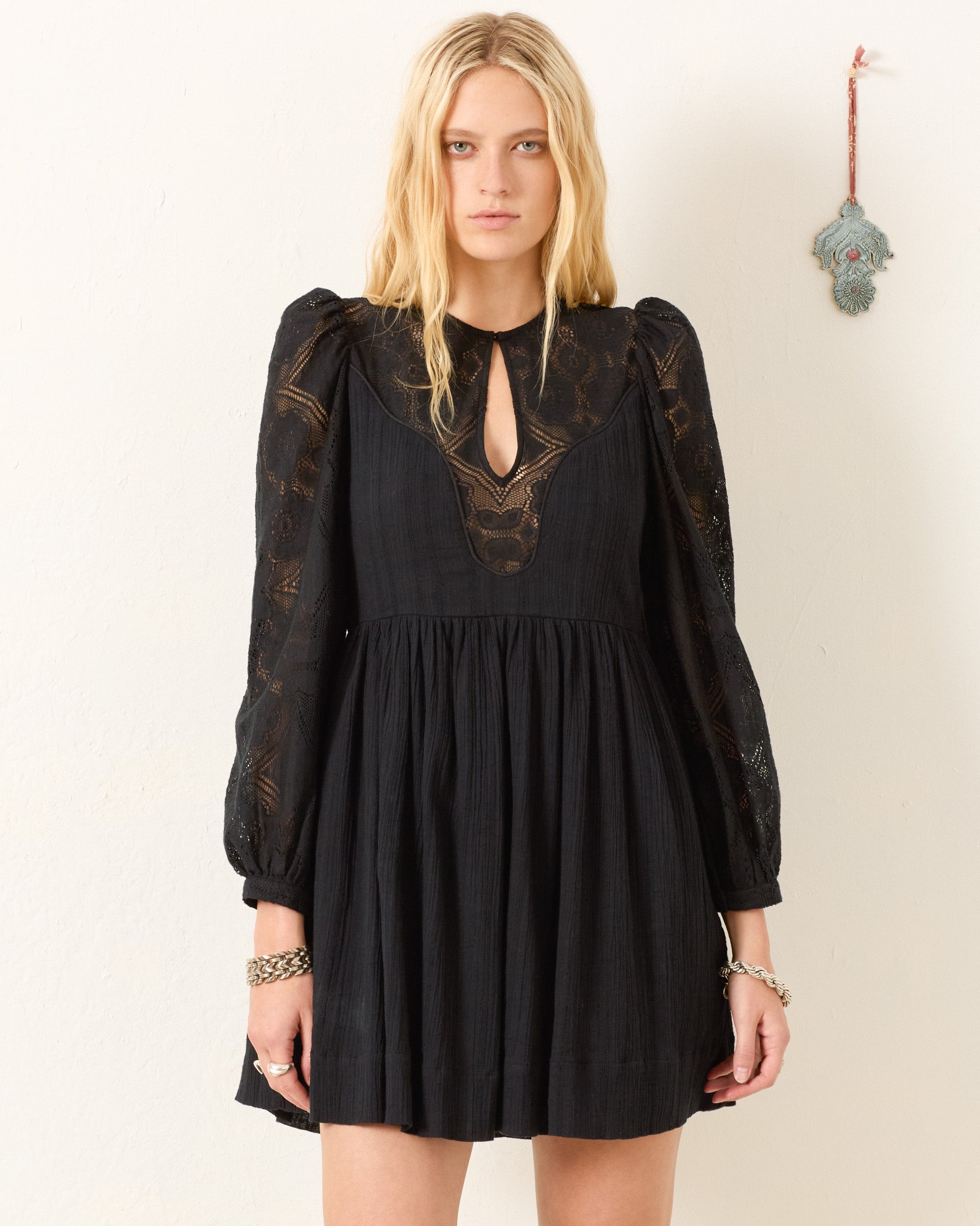 A woman wears the Winslow Shadow Lace Dress, a black mini dress with floral lace details, standing against a light wall with a small decorative item—ideal for holiday parties.