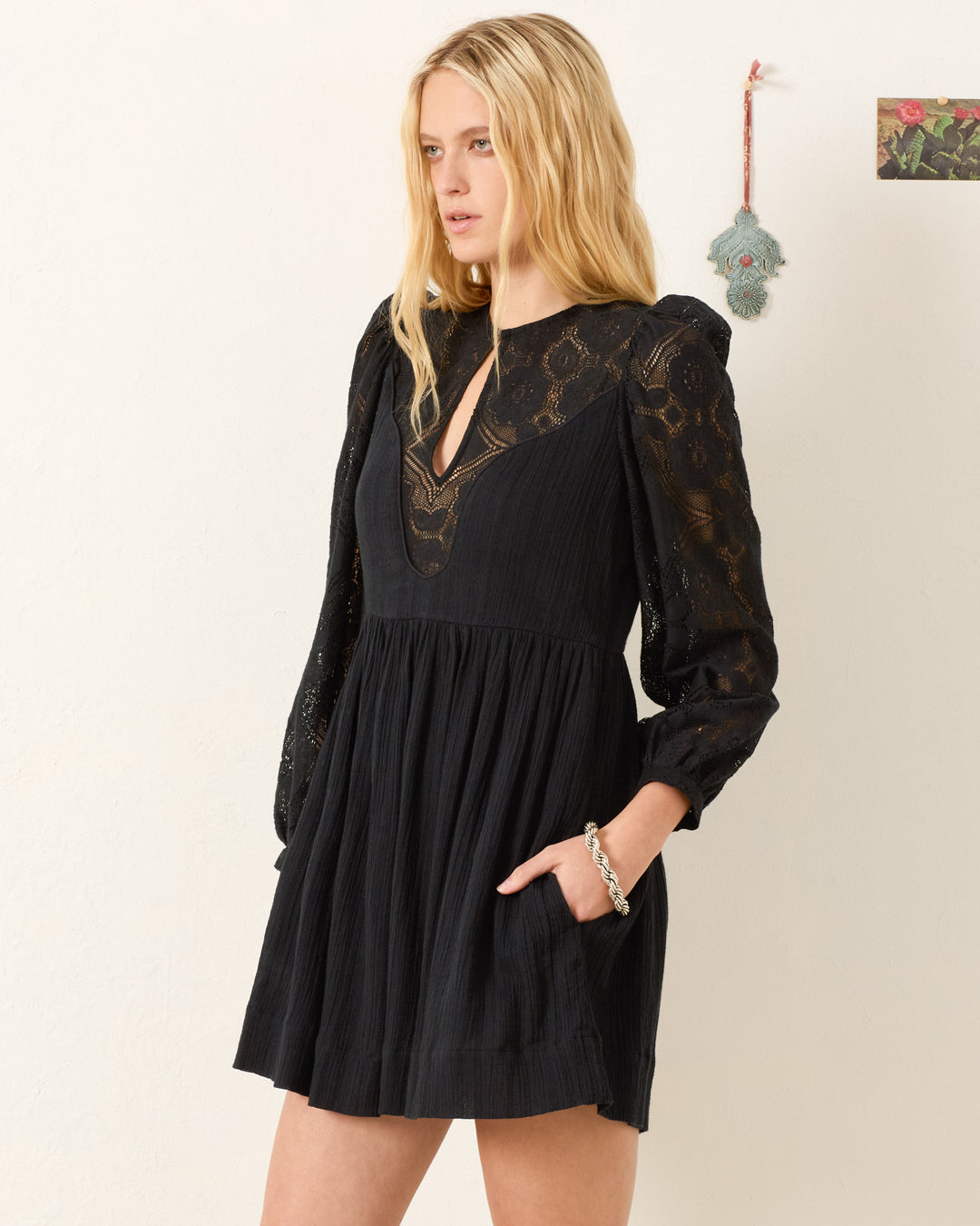 A woman with long blonde hair models the Winslow Shadow Lace Dress—a black mini dress with floral lace sleeves and a V-neckline. She stands against a light wall, hand in pocket—ideal for holiday parties.