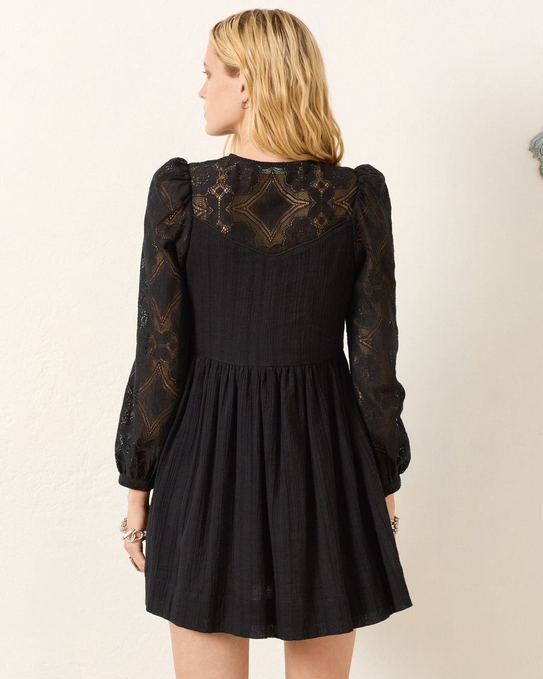 Woman stands with her back to the camera, wearing the Winslow Shadow Lace Dress—a black long-sleeve mini dress featuring elegant floral lace on the upper back and sleeves, perfect for holiday parties.