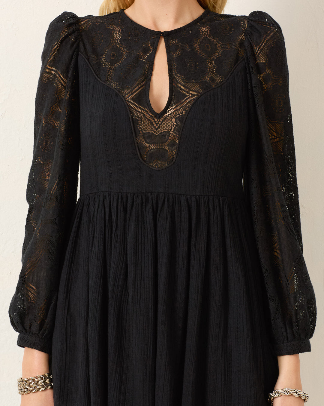 The Winslow Shadow Lace Dress is a black mini dress with floral lace on the chest, sleeves, and shoulders, a keyhole neckline, and long gathered sleeves—ideal for holiday parties.