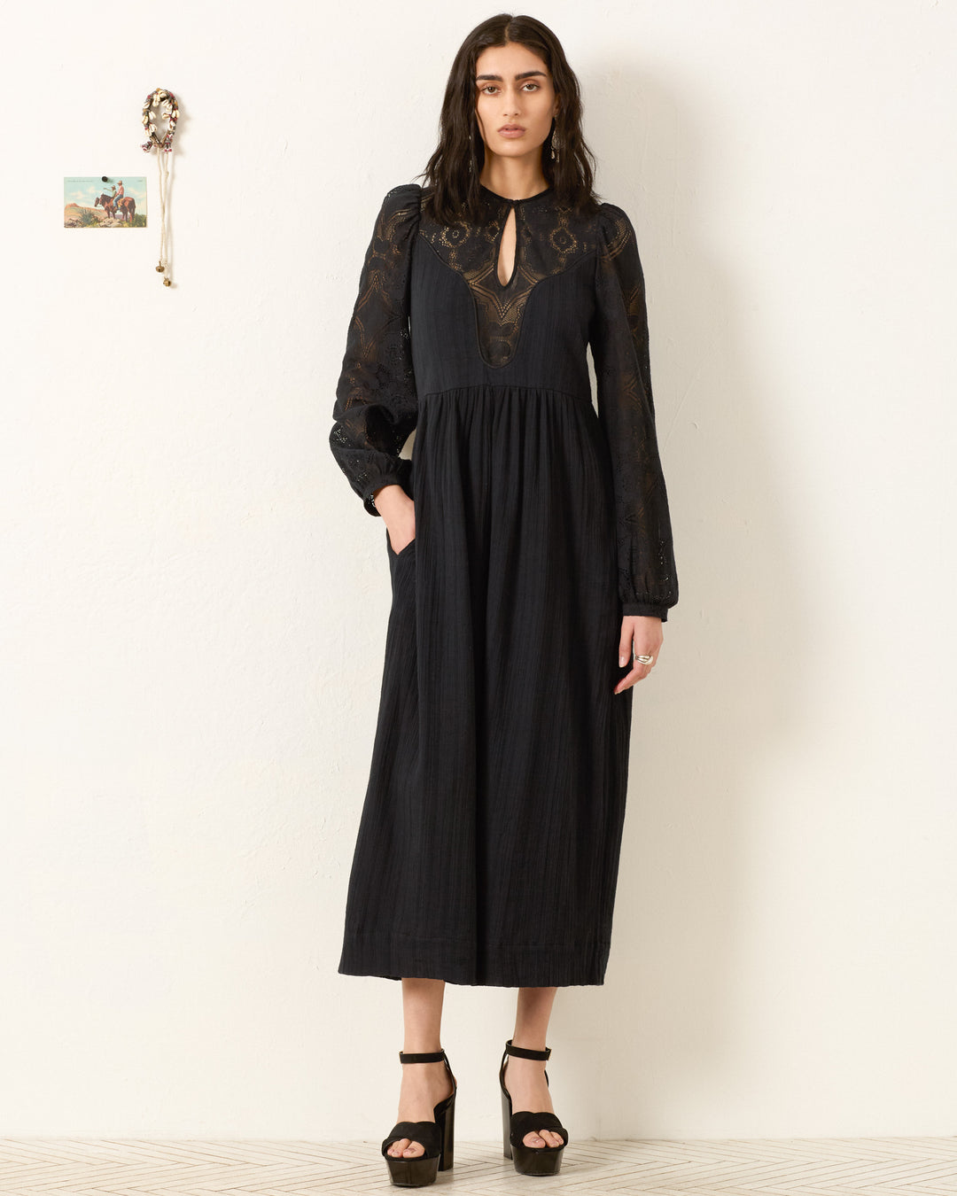 A woman stands against a white wall wearing the Winifred Shadow Lace Dress—its black maxi silhouette and floral lace sleeves paired with black high-heeled sandals make it an elegant choice for holiday parties.