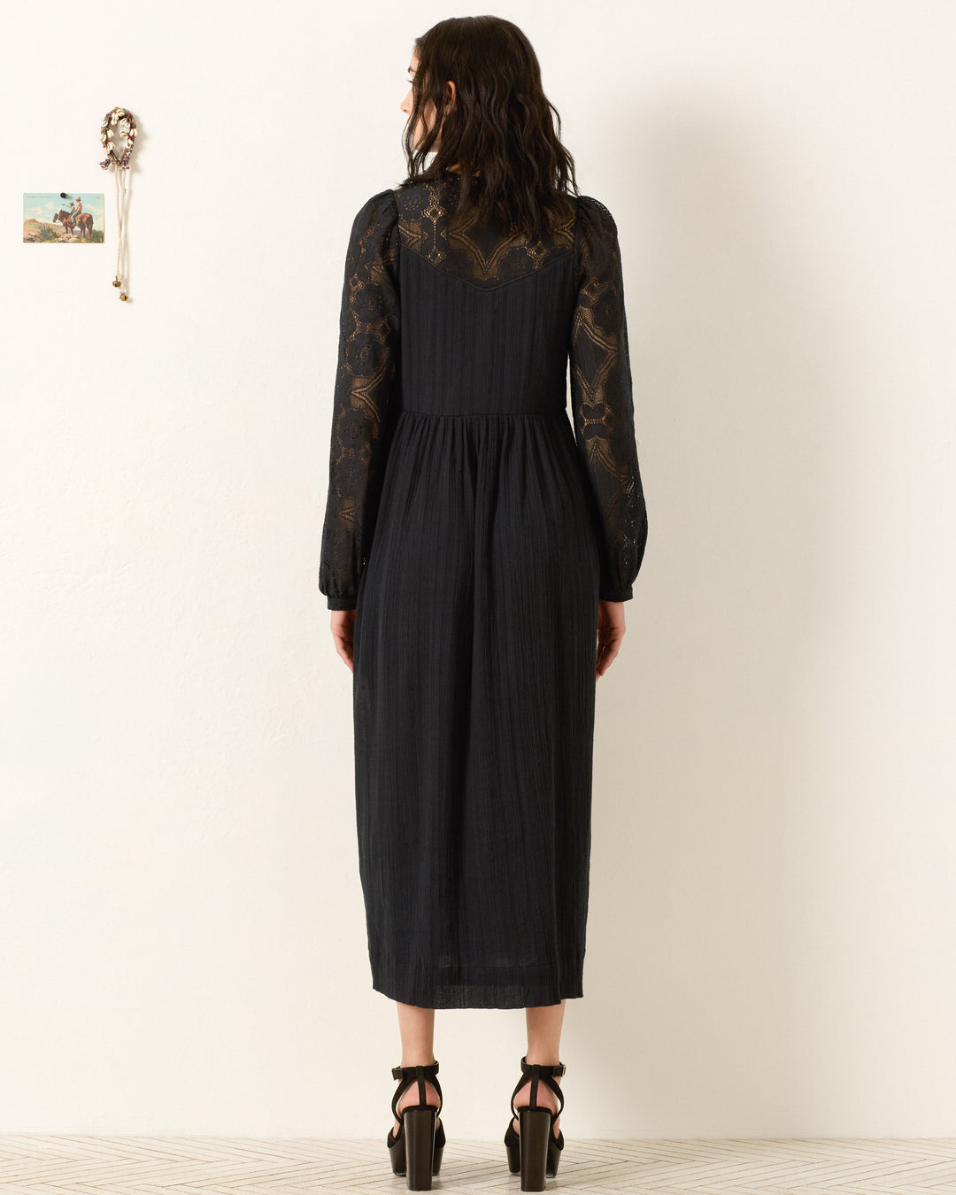 A woman faces a white wall wearing the Winifred Shadow Lace Dress, a long-sleeved black floral lace maxi—ideal for holiday parties. She pairs it with black heels, her wavy shoulder-length hair framing her look. Two wall decorations appear on the left.