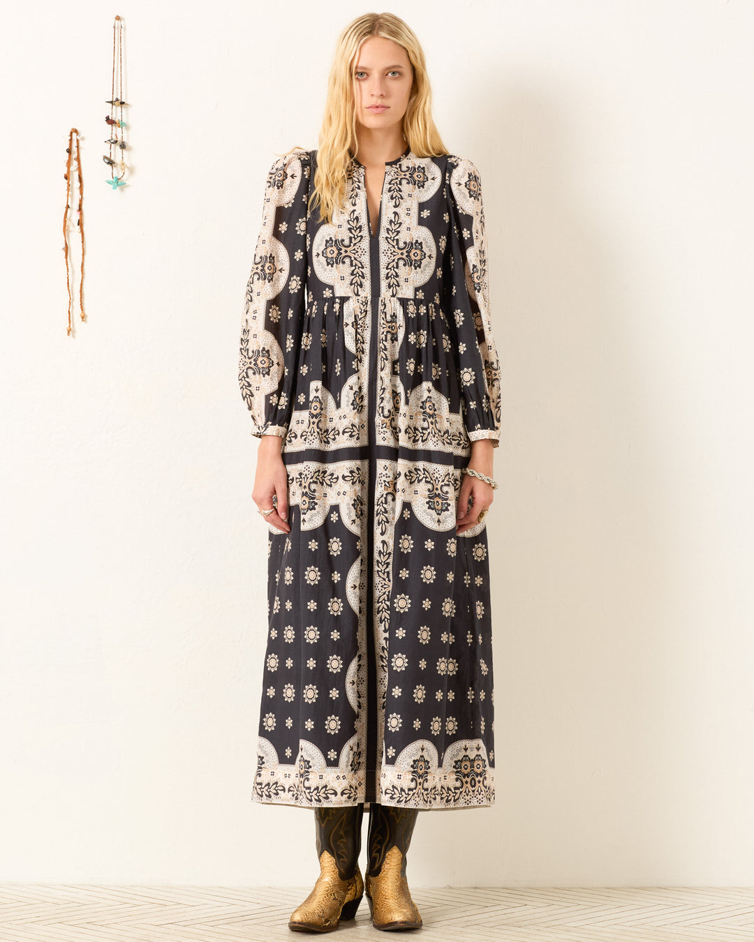 A woman stands against a white wall wearing the Winifred Bronco Bandana Dress—a Western-inspired, cotton-silk blend style with puffed sleeves—paired with gold cowboy boots; simple wall decor in the background.