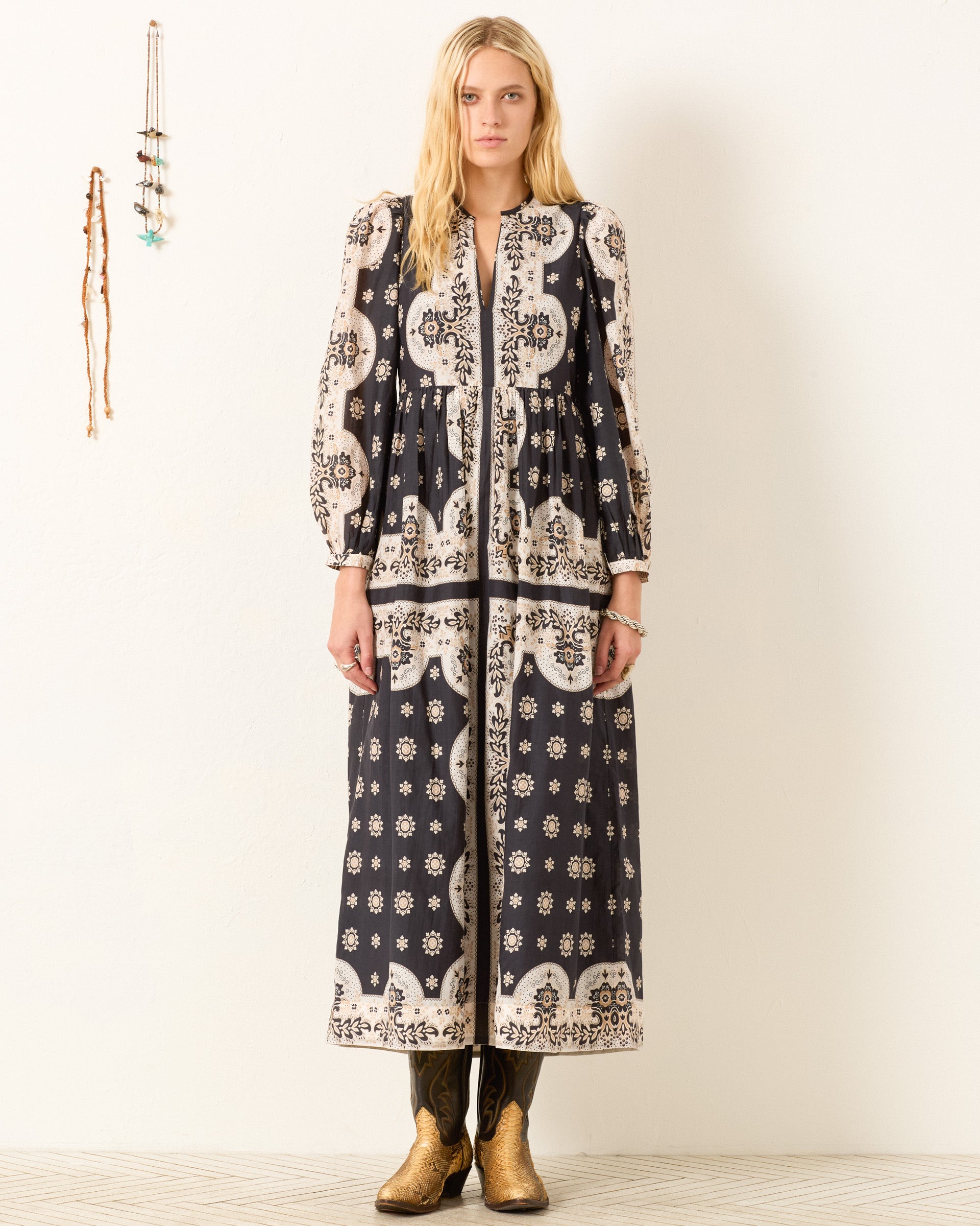 A woman stands against a white wall wearing the Winifred Bronco Bandana Dress—a Western-inspired, cotton-silk blend style with puffed sleeves—paired with gold cowboy boots; simple wall decor in the background.