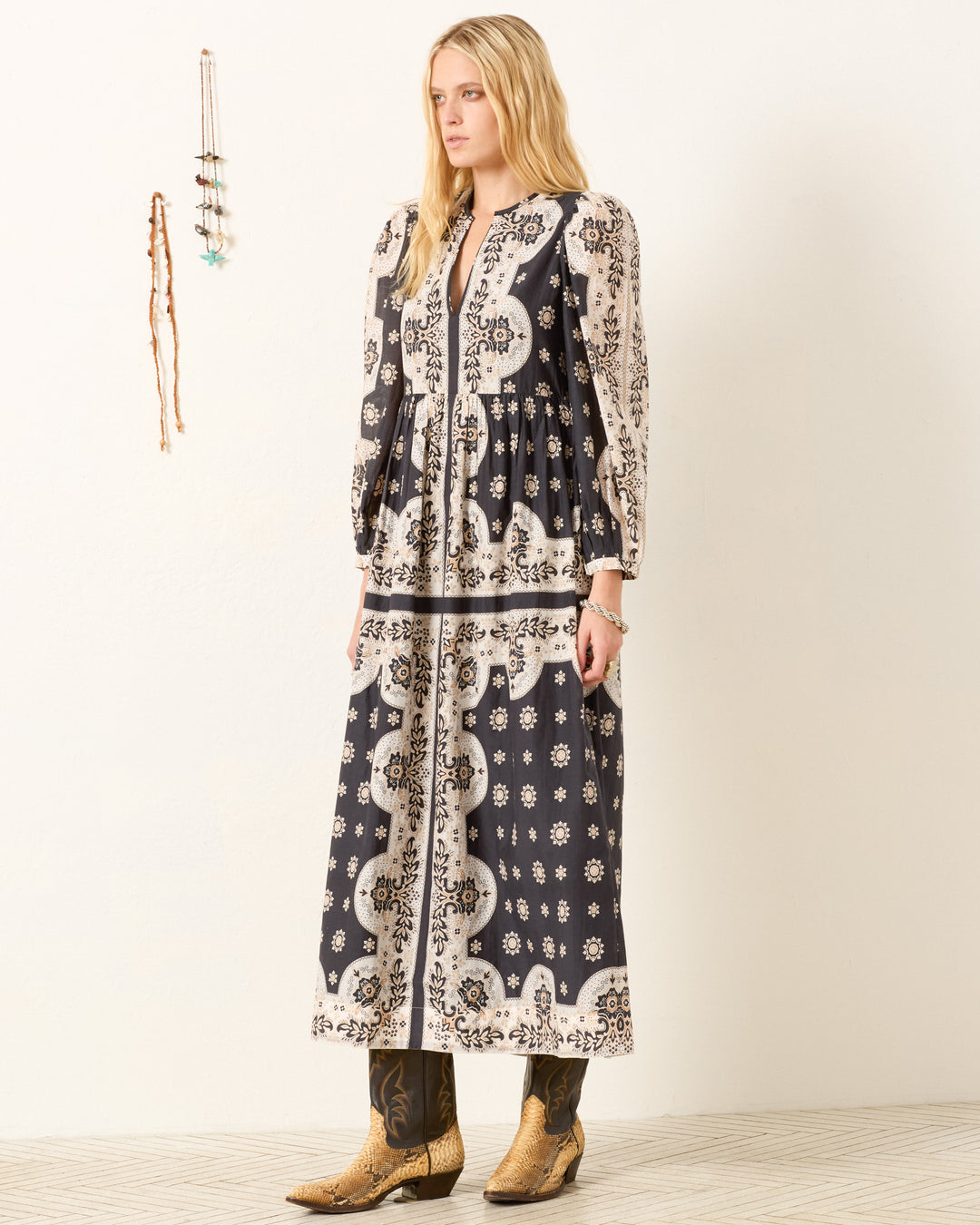 A woman stands indoors wearing the Winifred Bronco Bandana Dress with long sleeves and cowboy boots, facing left against a plain wall decorated with hanging jewelry.