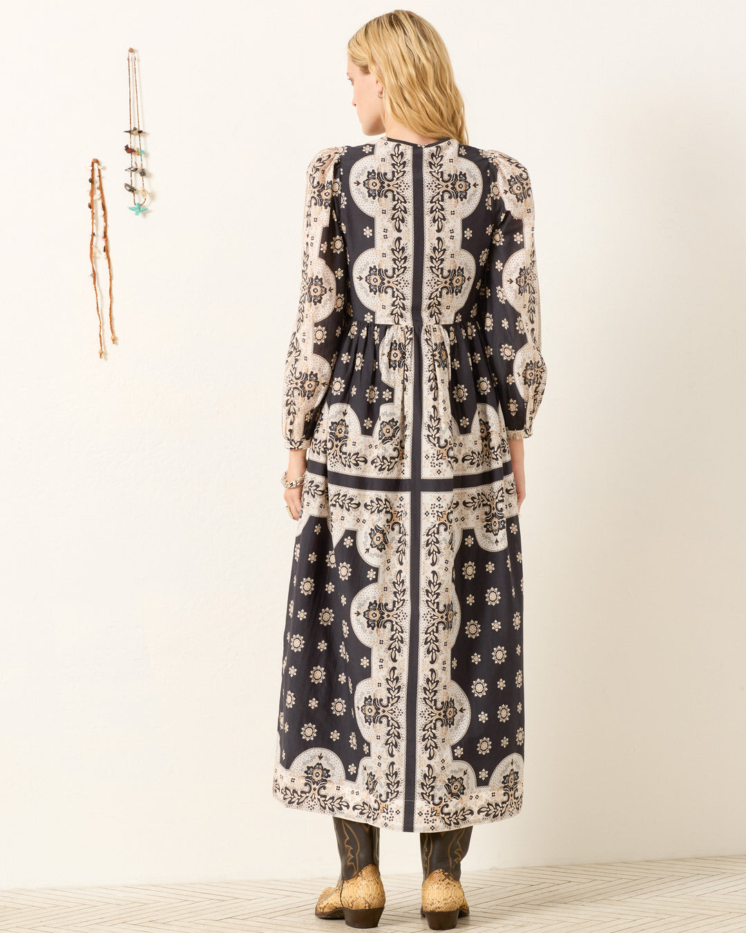 A woman faces away wearing the Winifred Bronco Bandana Dress, a long-sleeve, Western-inspired maxi with black and white patterns. She pairs it with tan boots and long earrings; necklaces hang on the wall beside her.