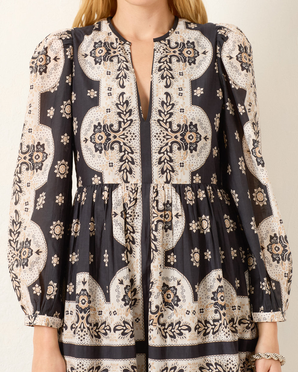 A woman wearing the Winifred Bronco Bandana Dress, a long-sleeved, Western-inspired style with black and beige floral and geometric prints, gathered waist, and a front slit neckline.
