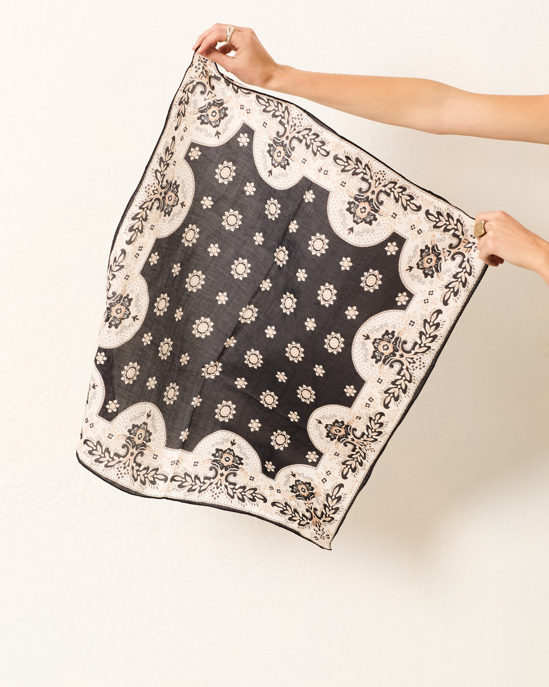 A person holds up the Beau Bronco Bandana, a square black cotton-silk accessory with a white floral and decorative border, showcasing its vintage-inspired style against a plain light background.