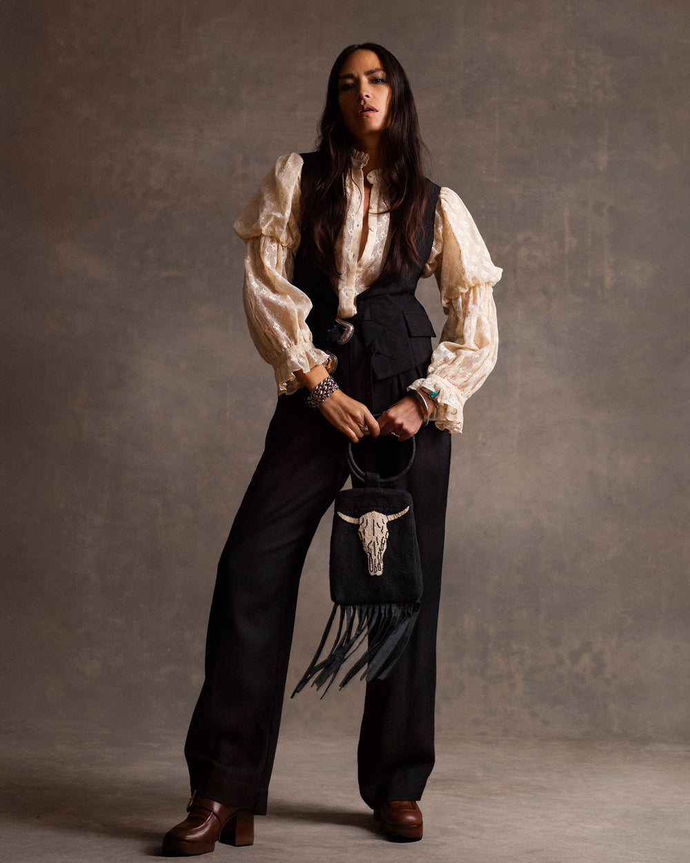 A person stands against a neutral backdrop wearing the Mazie Antique Bone Lace Shirt, black vest and trousers, brown platform shoes, and holding a fringed black bag with a decorative skull for a ’70s dandy style touch.