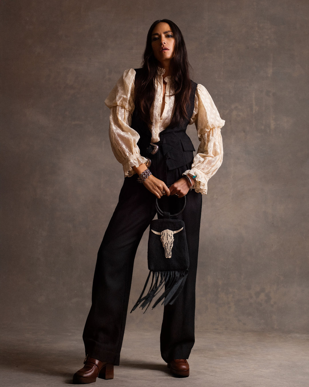 A person stands against a neutral backdrop wearing the Mazie Antique Bone Lace Shirt, black vest and trousers, brown platform shoes, and holding a fringed black bag with a decorative skull for a ’70s dandy style touch.