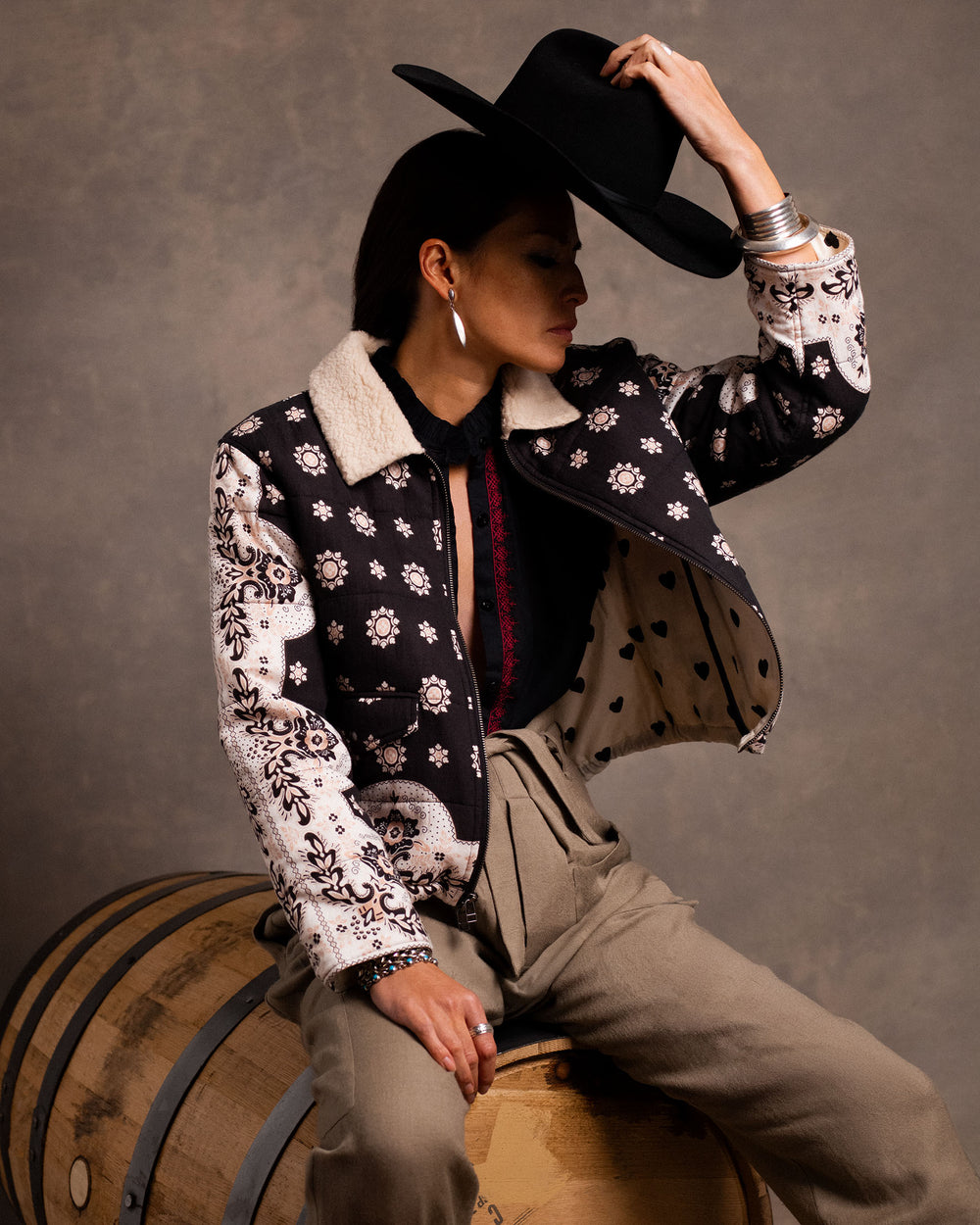 A person sits on a barrel wearing the Lex Bronco Bandana Jacket, high-waisted pants, and holding a black hat, set against a neutral background.