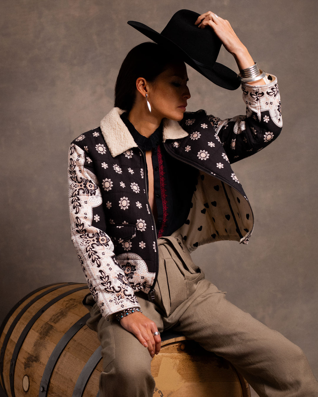 A person sits on a barrel wearing the Lex Bronco Bandana Jacket, high-waisted pants, and holding a black hat, set against a neutral background.