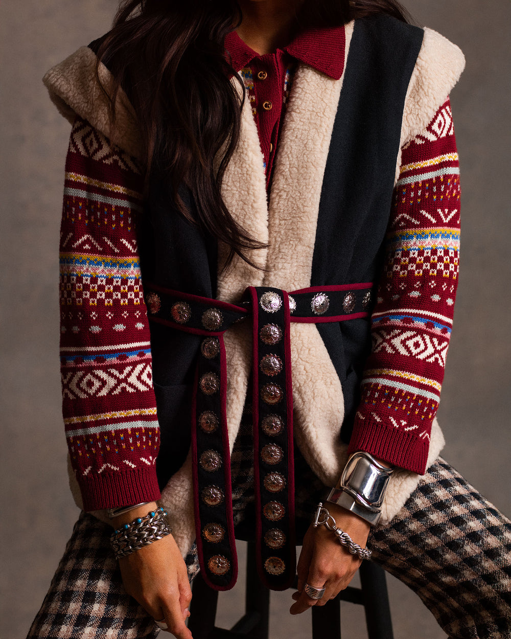 Wearing the Liza Mesa Fair Isle Top, paired with a black and beige sleeveless vest, studded belt, plaid pants, and silver bracelets, a person sits on a stool.