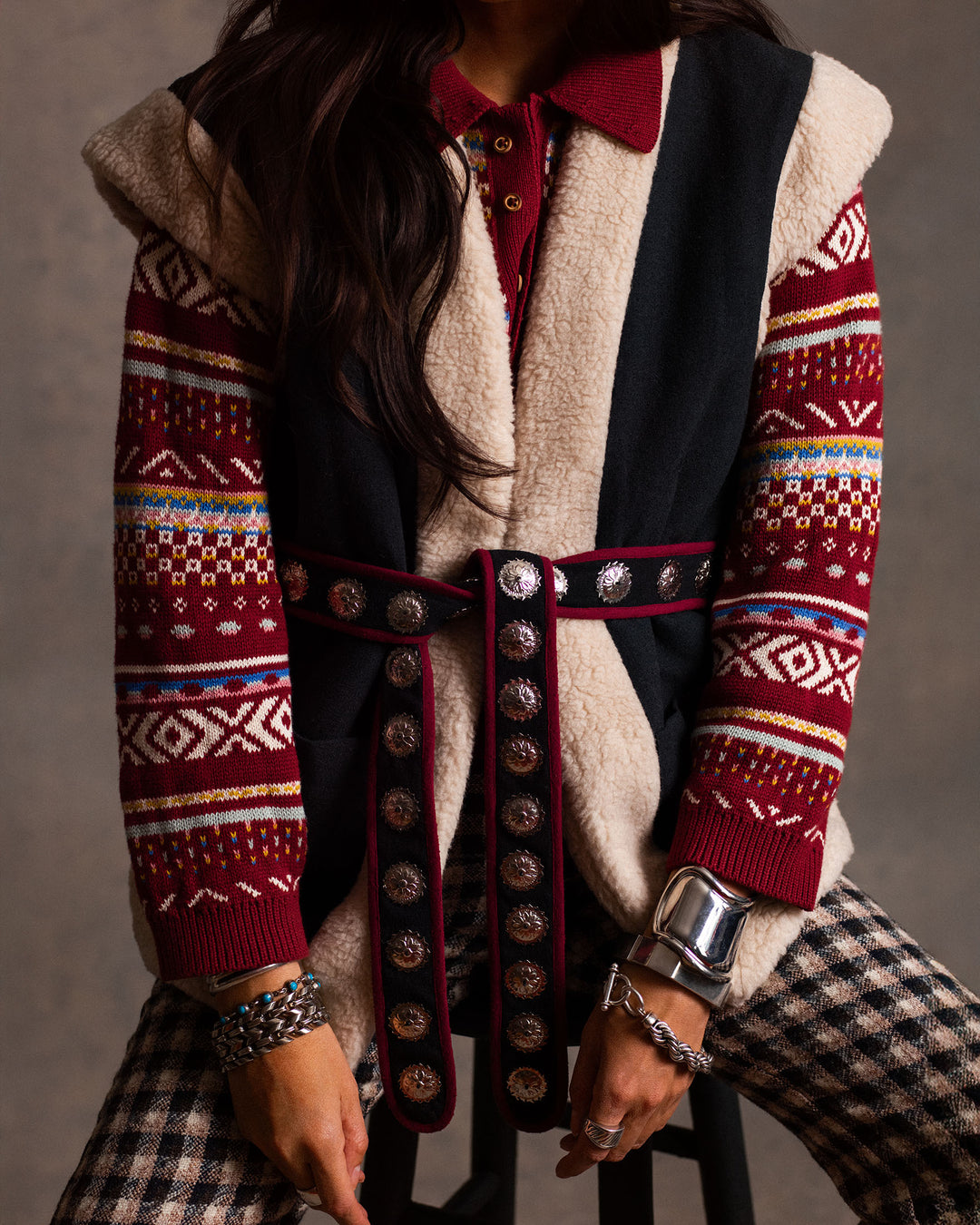 Wearing the Liza Mesa Fair Isle Top, paired with a black and beige sleeveless vest, studded belt, plaid pants, and silver bracelets, a person sits on a stool.