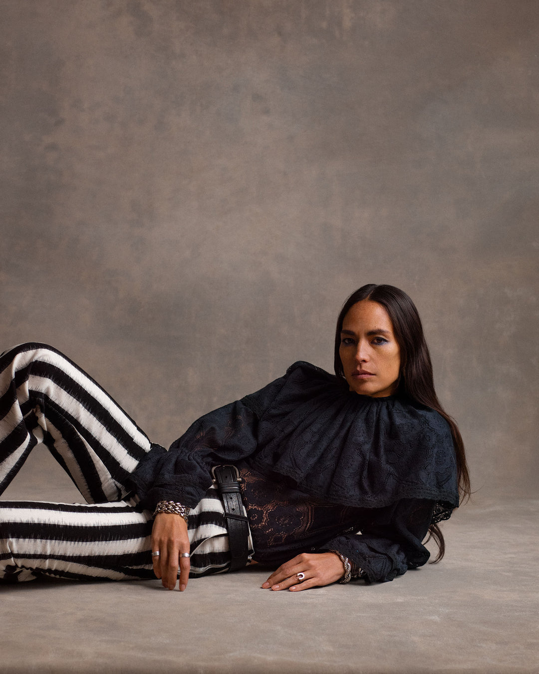 A person with long dark hair reclines on the floor, wearing the Eloise Shadow Lace Top with Victorian sleeves and black-and-white striped pants against a neutral studio backdrop.