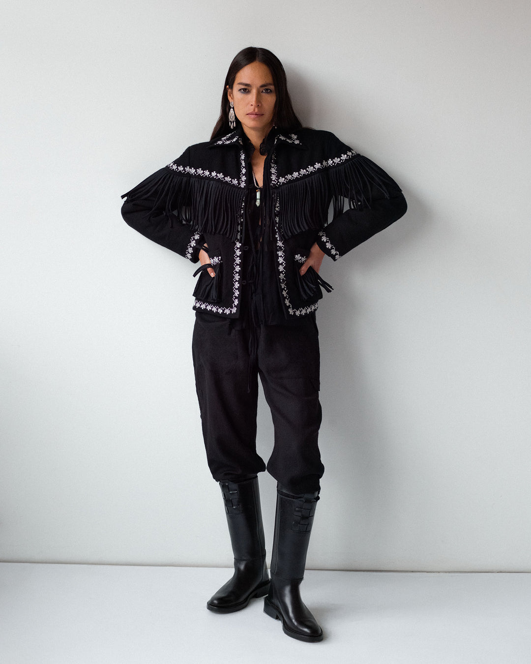 A woman stands against a white wall wearing the Diana Noir Wool Pant, styled with a black fringed jacket with white trim and black knee-high boots, hands resting on her hips.