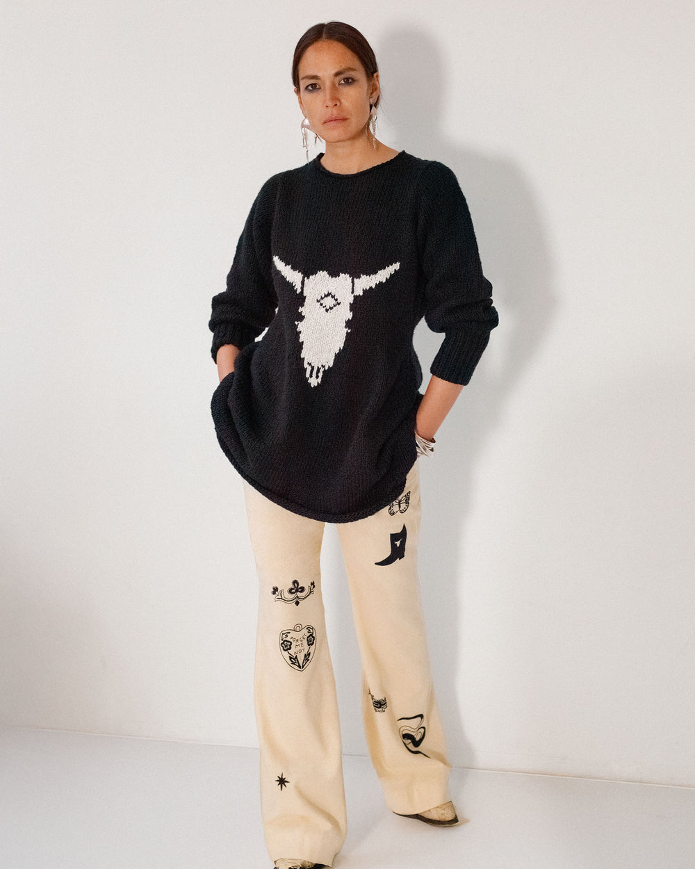 A woman stands against a white wall wearing the Renny Cow Skull Dress, featuring an organic cotton fabric with a white bull skull graphic and cream fabric decorated with various black symbols.