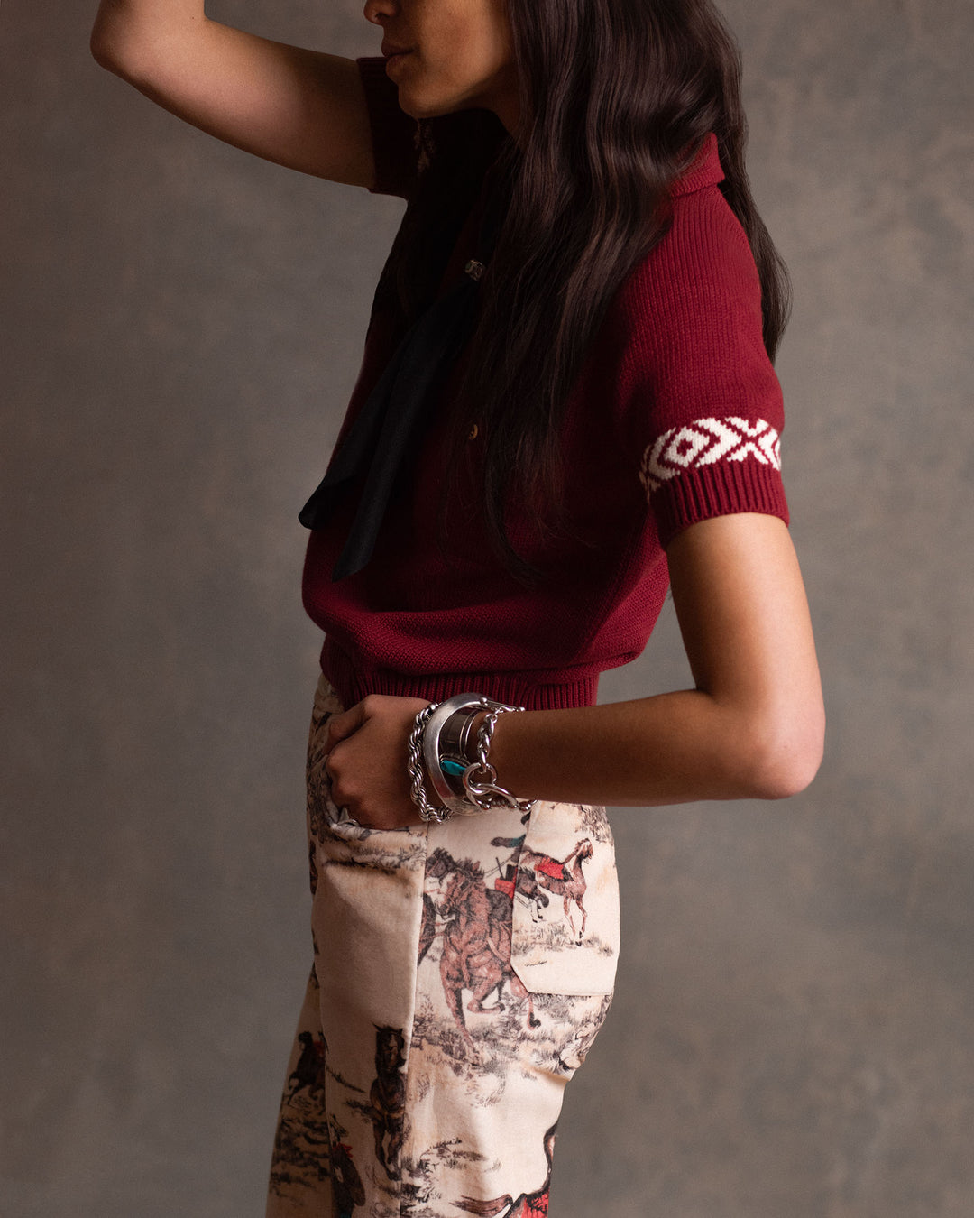 A woman in profile models the Bunny Canyon Cowboy Jean, featuring a flared leg and cowboy print, paired with a maroon short-sleeve sweater and multiple bracelets against a neutral background.