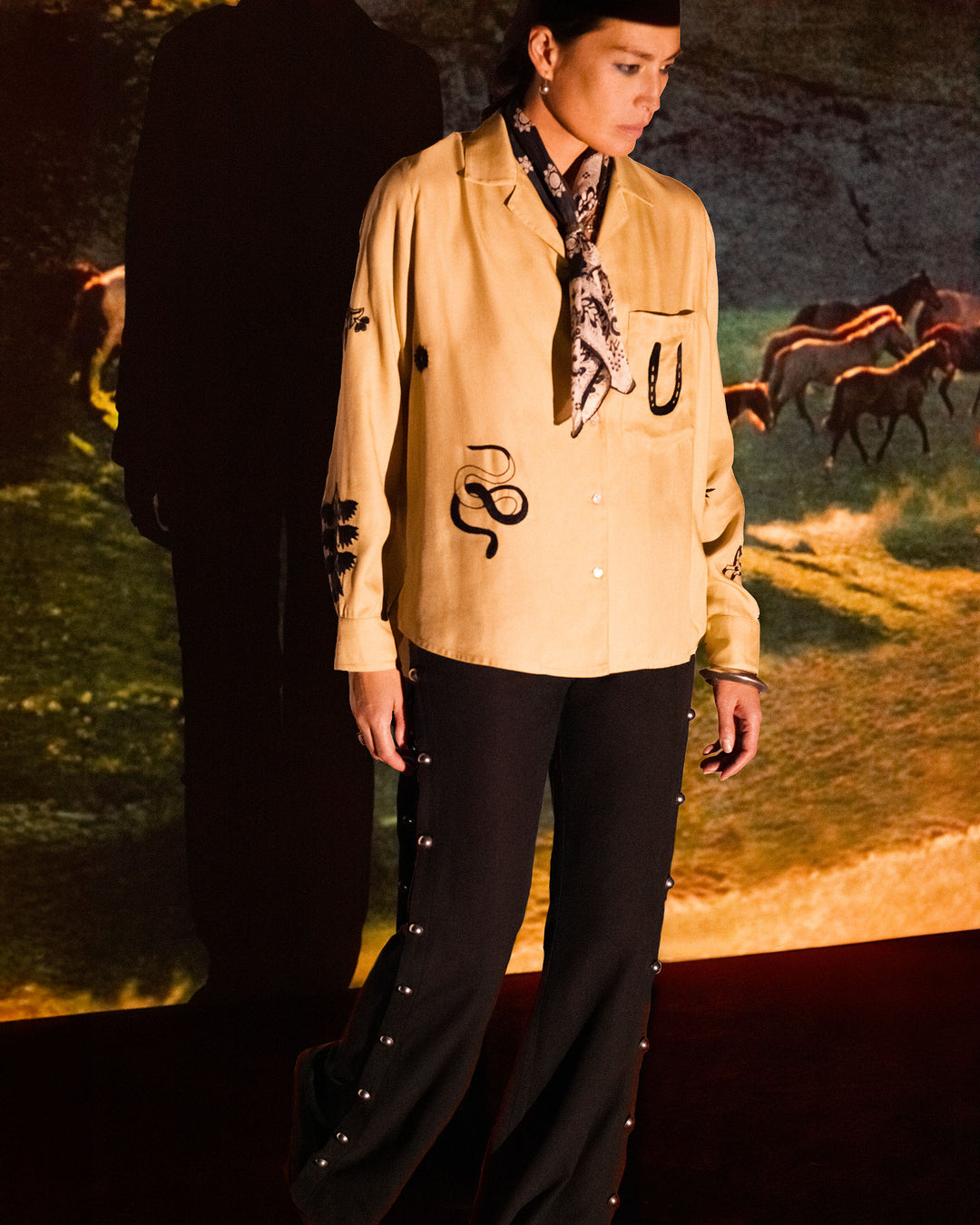 A person wearing the Patti Desert Tattoo Shirt— a beige button-up with black snake designs—black pants, and a patterned scarf stands in front of a projected image of running horses.