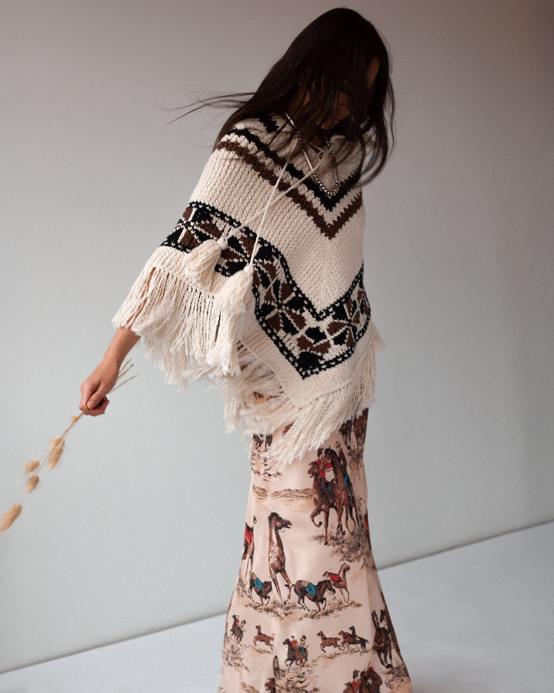 A person wears the Faune Rawhide Knit Poncho, featuring fringed edges and geometric patterns, paired with a long skirt with a horse and cowboy print, standing against a plain background.