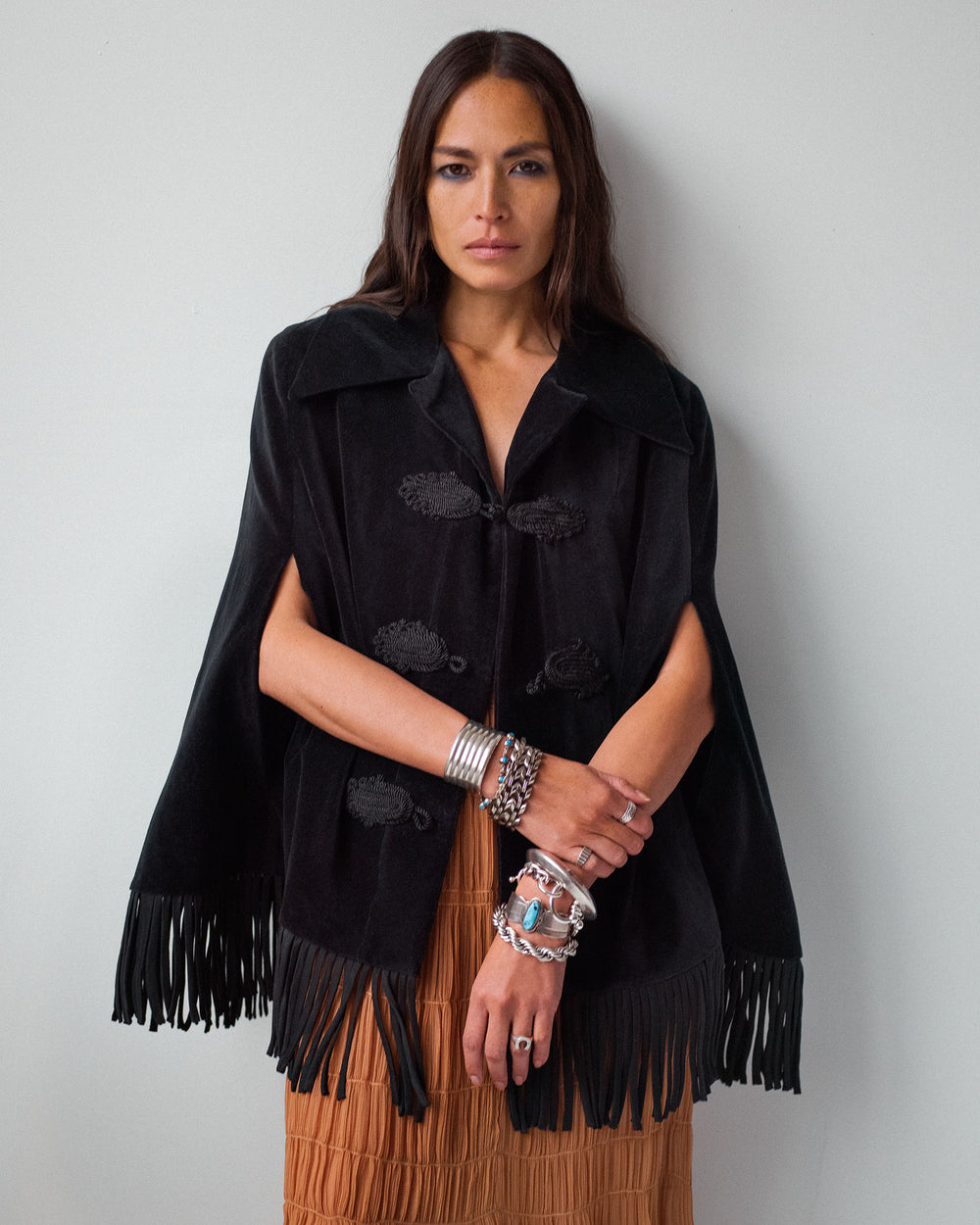 A woman with long brown hair models the Kensington Onyx Fringe Cape over a brown skirt, accessorized with silver bracelets and rings, standing against a plain light-colored wall.