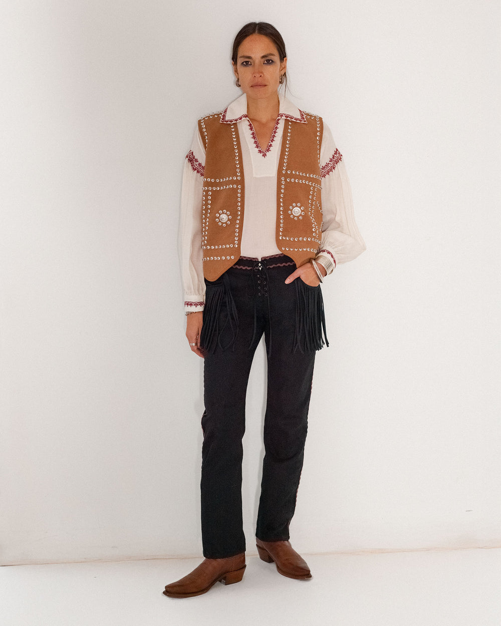 A woman stands against a white wall wearing a tan embellished vest over a white blouse, Ezra Noir Rein Pant with fringe, and brown boots.