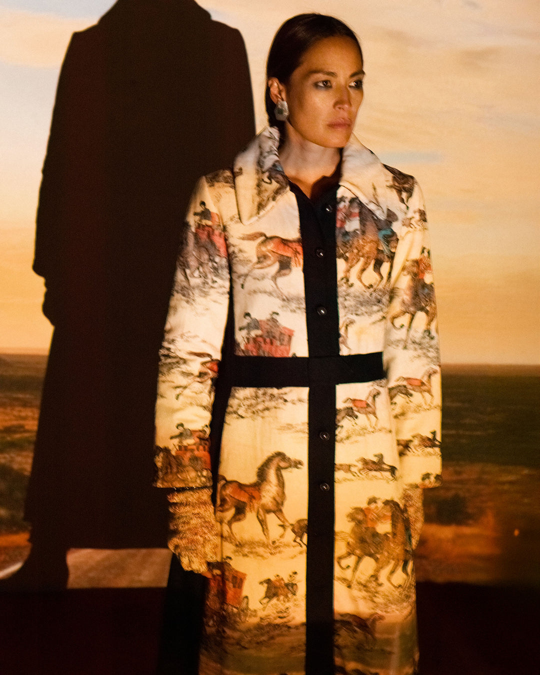 A woman in the Lara Canyon Cowboy Coat, featuring vintage horse and rider prints, stands before a projected cowboy desert scene with her shadow cast behind her.