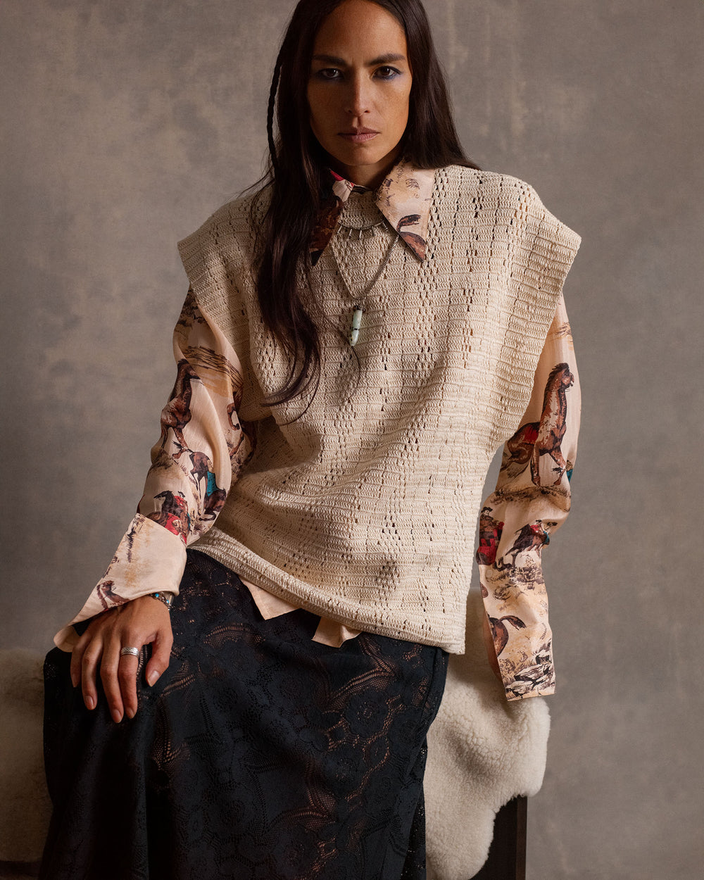 A woman sits on a bench wearing the Dana Canyon Cowboy Silk Shirt under a beige sleeveless knit sweater and paired with a dark lace skirt, set against a neutral background.