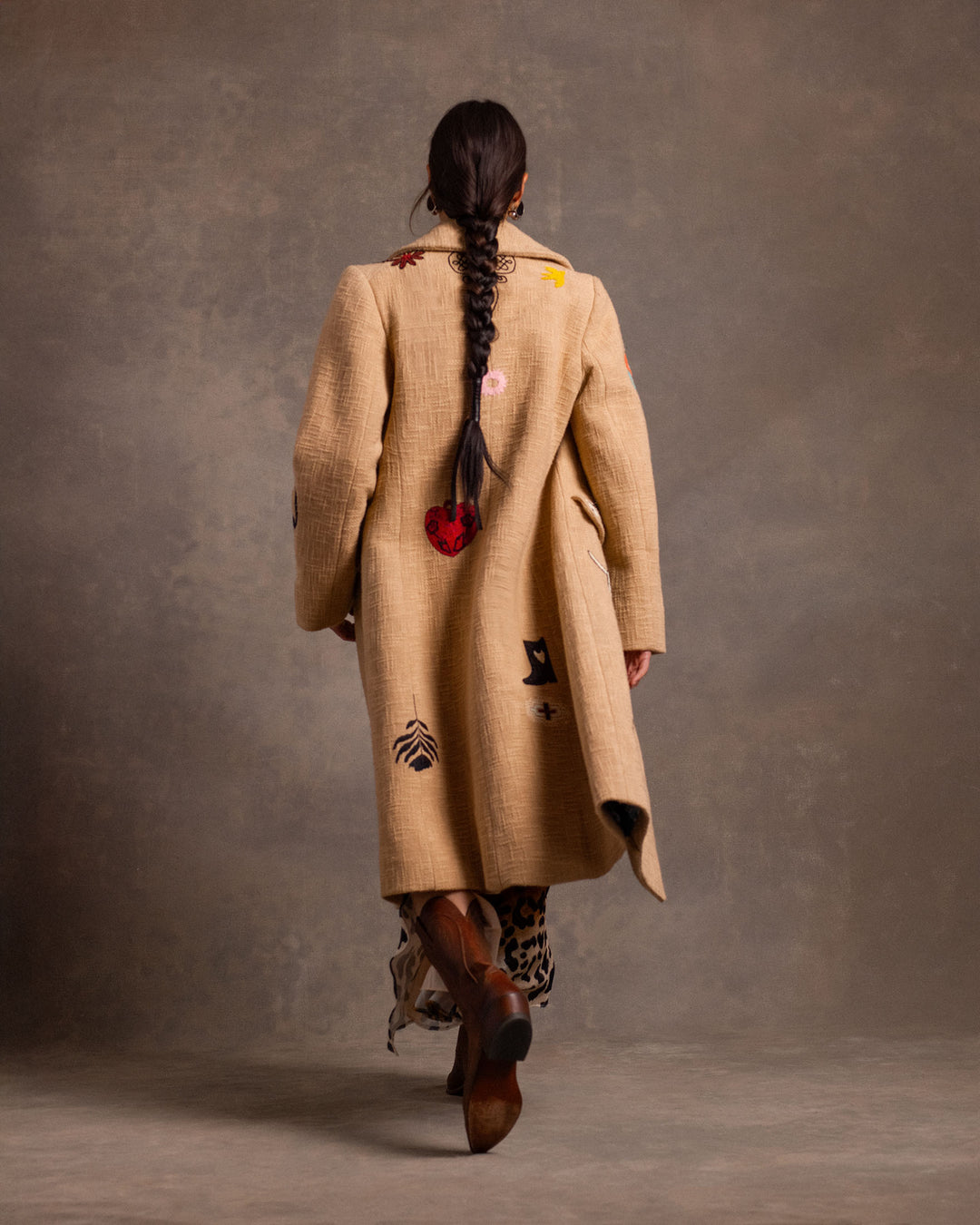 A person with long braided hair wearing the Petunia Desert Tattoo Coat—Western-inspired and beige with colorful patches—and heeled boots, walks away from the camera against a plain brown backdrop.