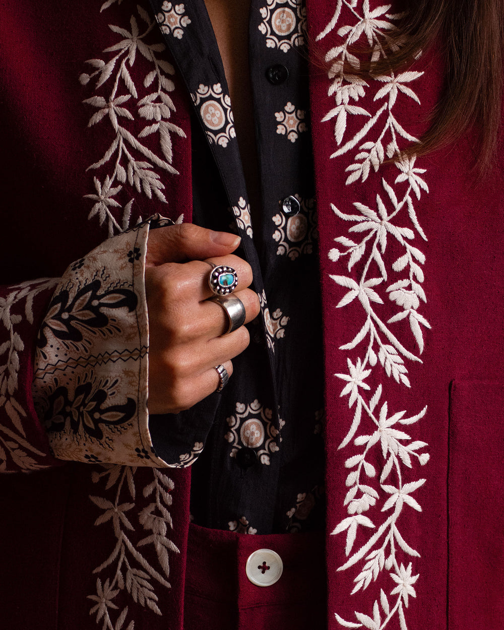 A person wearing the Dana Bronco Bandana Shirt and a maroon jacket with white floral embroidery, displaying rings on their fingers.