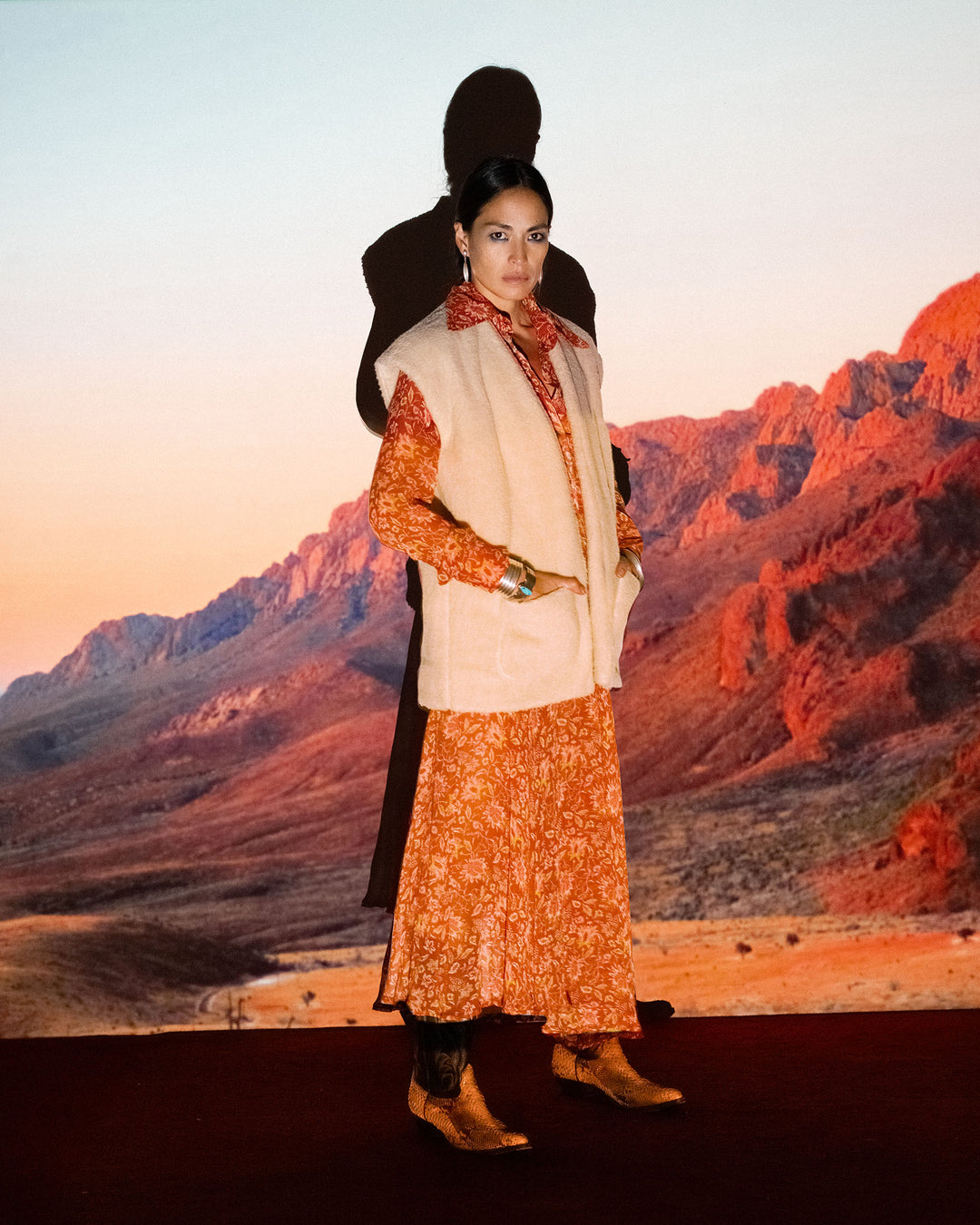 A woman stands on a red carpet before a red mountain backdrop, wearing a patterned orange dress, brown boots, and the Harrow Cactus Spur Vest. Her shadow is visible on the backdrop.