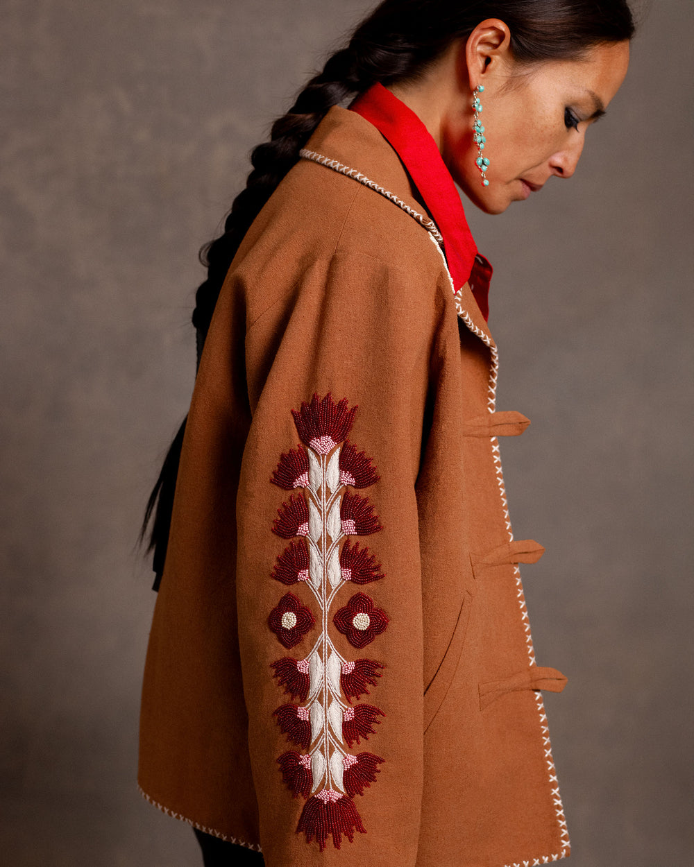 A woman with a long braid wears the Western Cactus Bloom Jacket, featuring red and white sleeve embroidery, over a red shirt against a neutral background.