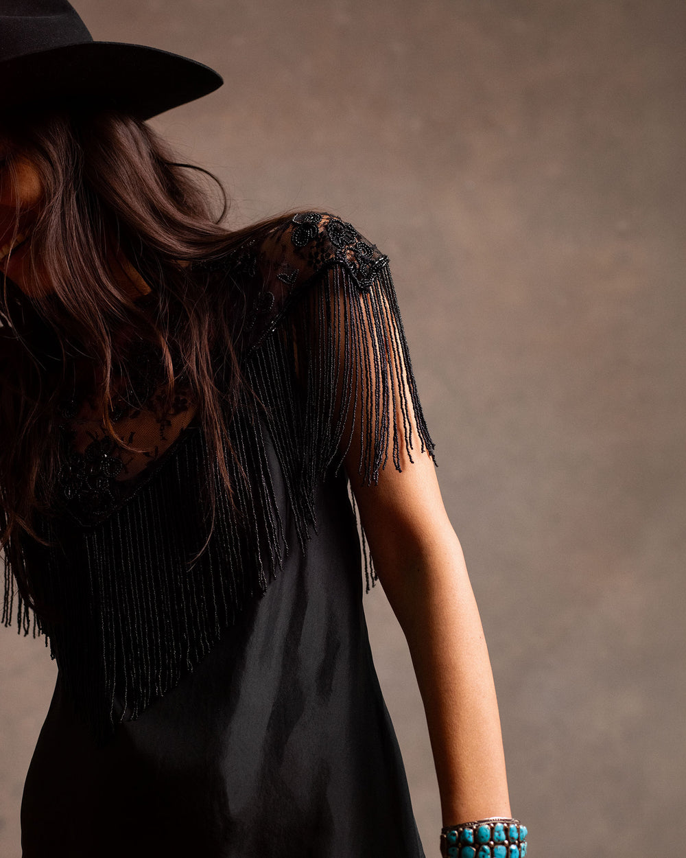 A woman in the Adele Noir Silk Dress and a black hat stands against a brown background, wearing a turquoise beaded bracelet on her wrist.